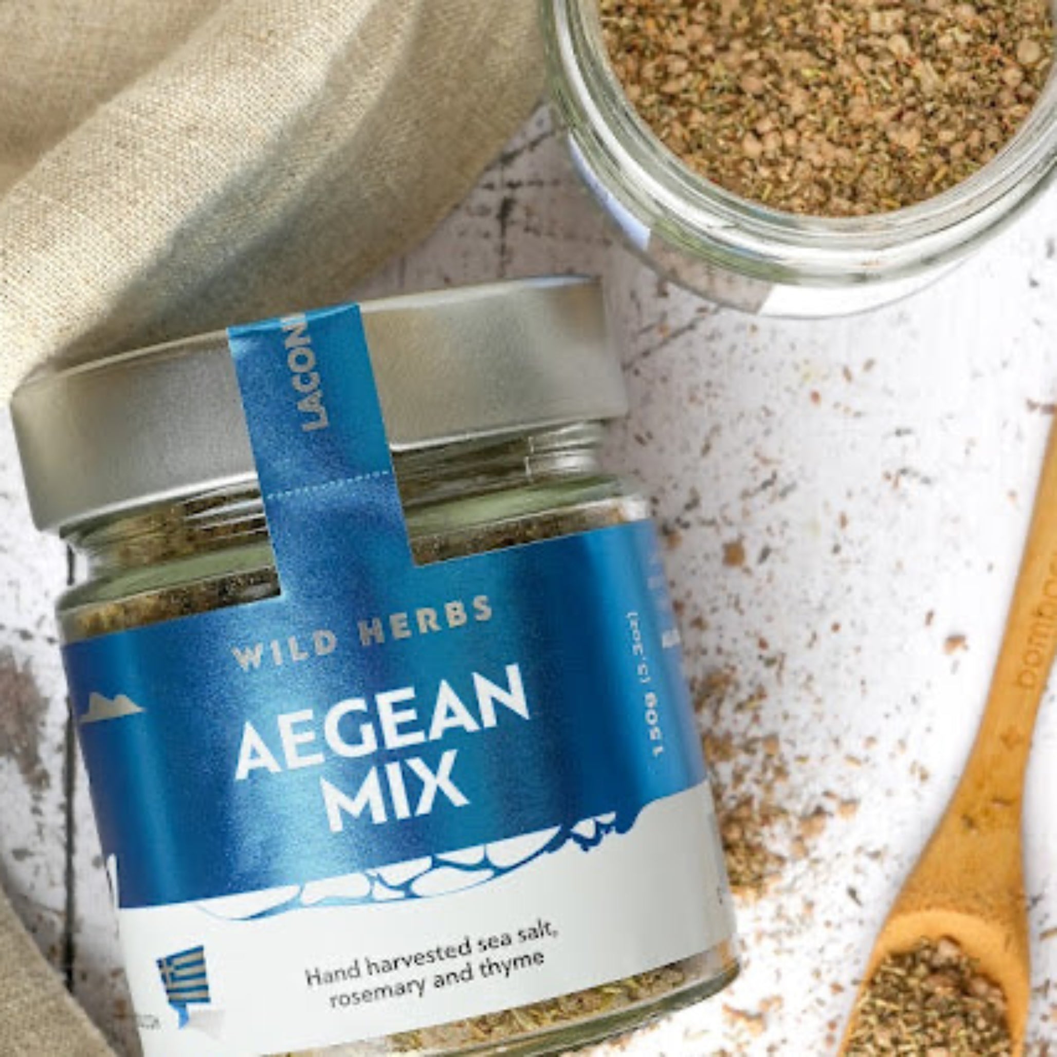 Aegean Mix Greek sea salt blend jar with wild rosemary and thyme, shown with wooden spoon and linen cloth, herbs scattered on white surface.