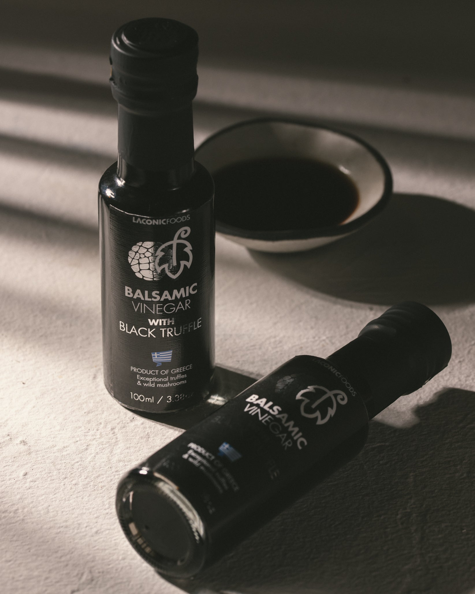 Two bottles of Laconic Foods Balsamic Vinegar with Black Truffle on a textured surface with dramatic shadows and a bowl of balsamic.