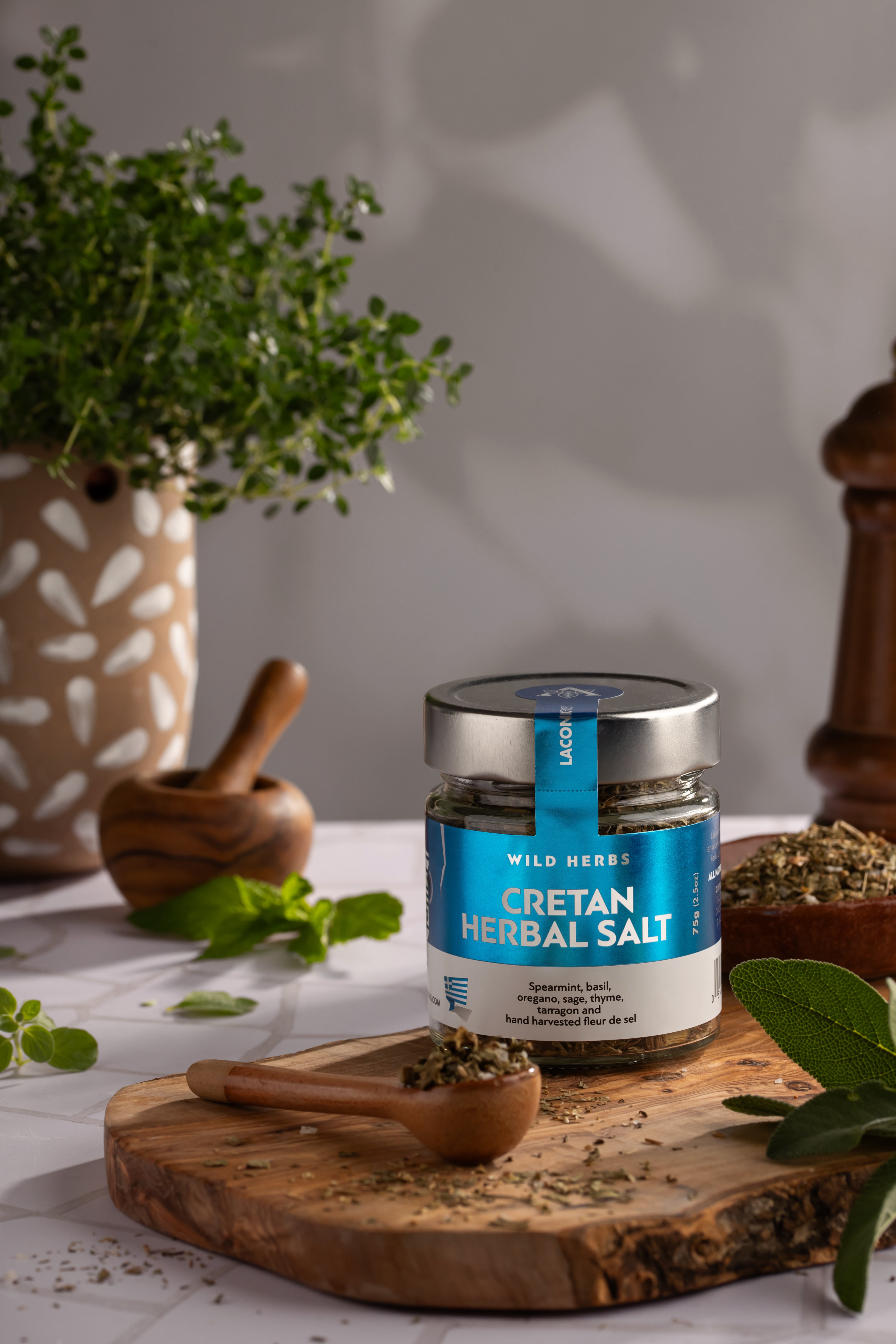 Cretan Herbal Salt by Laconic Foods on a wooden board, surrounded by Greek herbs and rustic kitchen tools in a Mediterranean setting