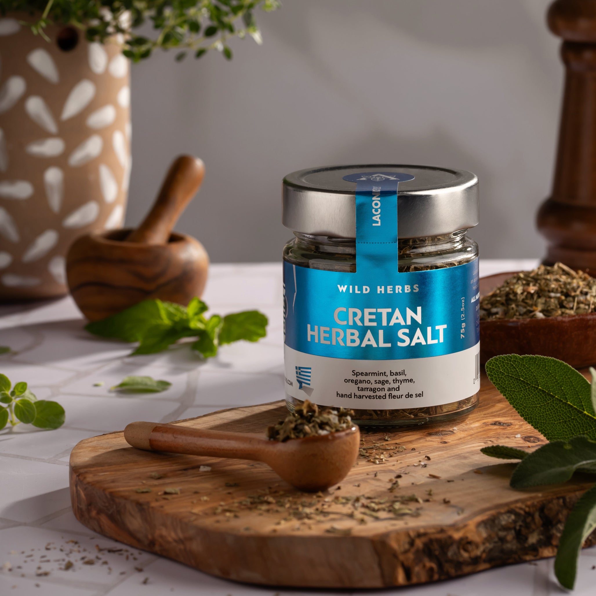 Cretan Herbal Salt by Laconic Foods on a wooden board, surrounded by Greek herbs and rustic kitchen tools in a Mediterranean setting