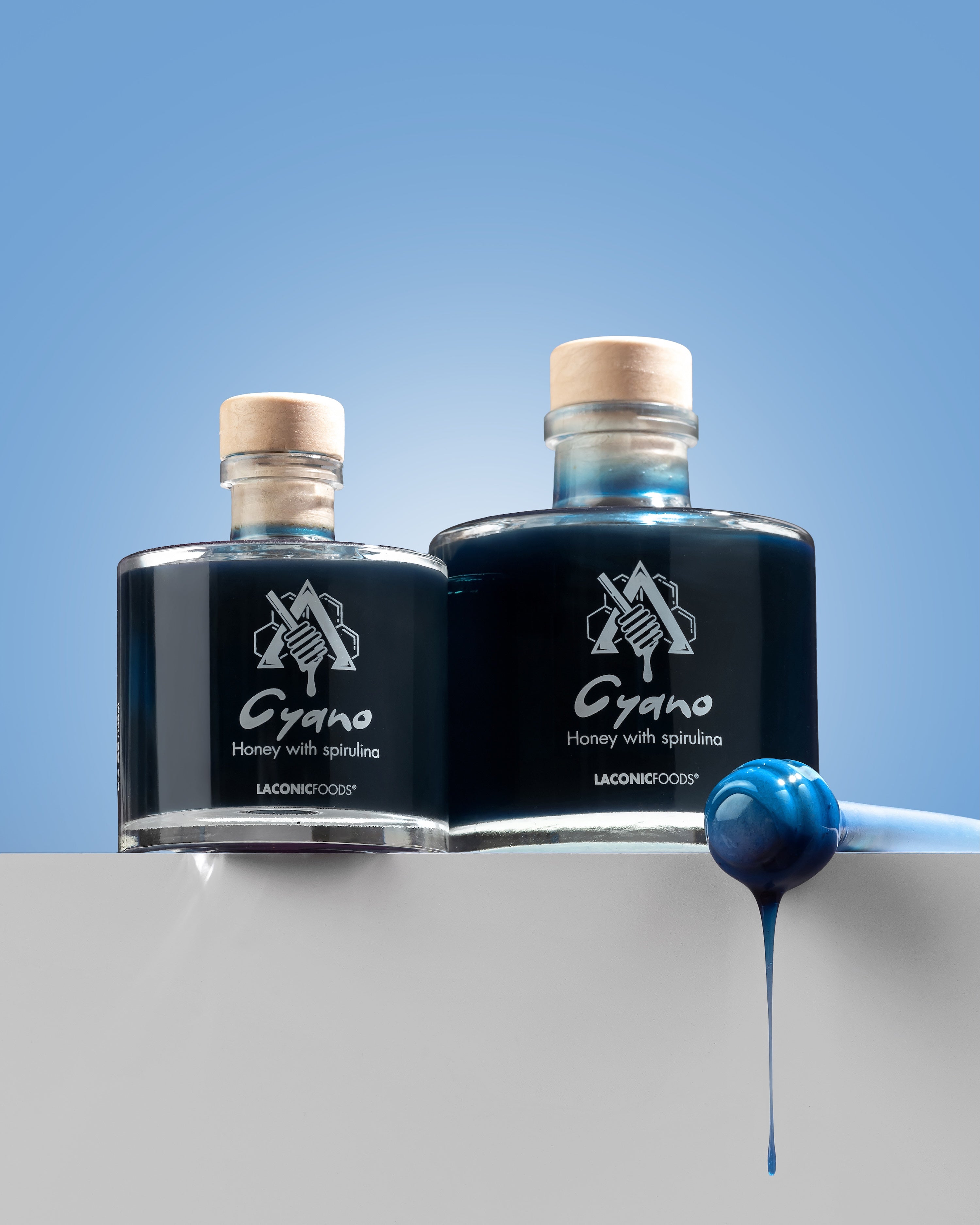 Two glass bottles of Cyano blue honey with spirulina and a honey dipper β Laconic Foods product shot.
