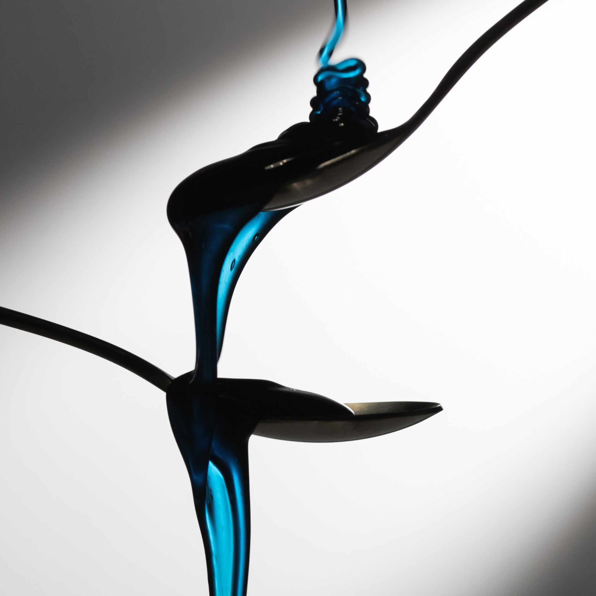 Dramatic pour of Cyano blue honey with spirulina over stacked spoons, highlighting the deep blue tone.