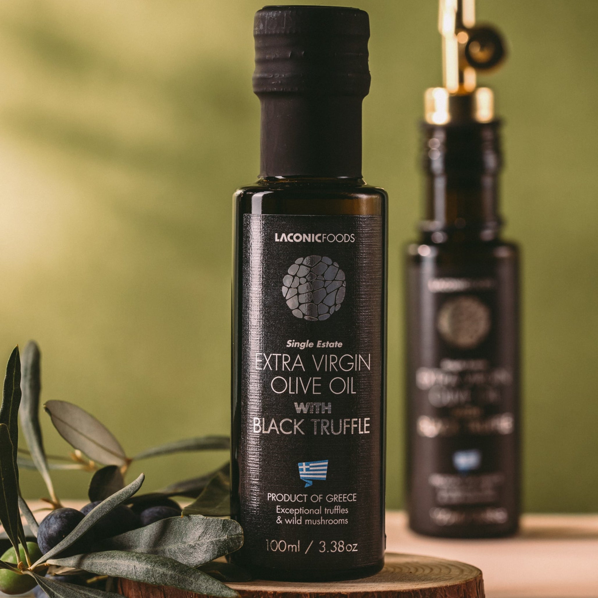 Bottle of Laconic Foods Extra Virgin Olive Oil with Black Truffle displayed on a wooden board with fresh olives and olive leaves.