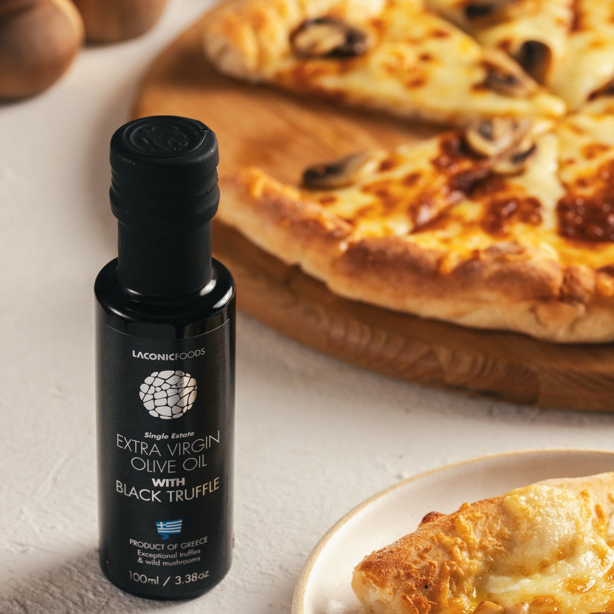 Laconic Foods Extra Virgin Olive Oil with Black Truffle placed beside a mushroom pizza slice on a plate and wooden pizza board.