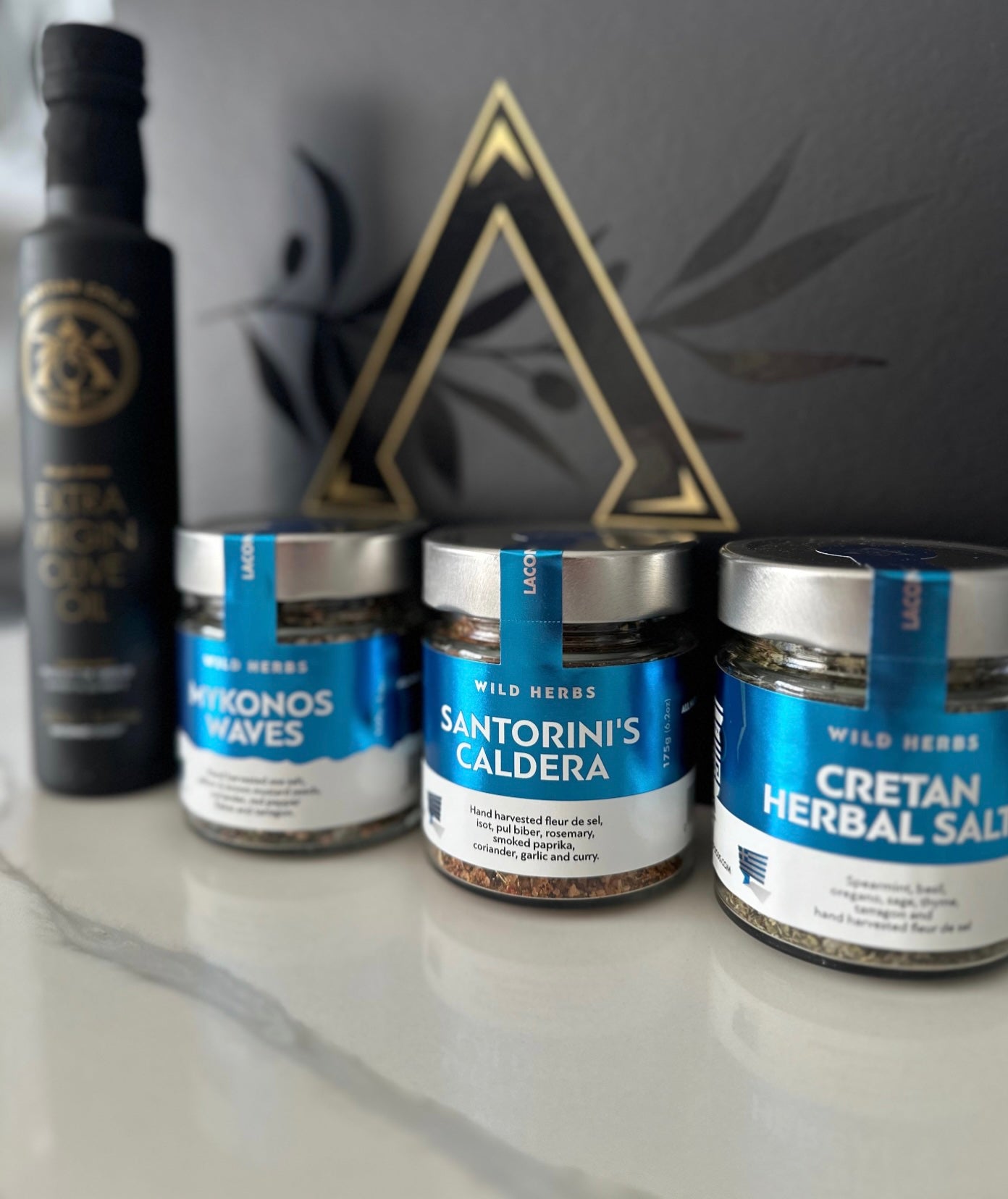Close-up of Laconic Foods Mykonos Waves, Santorini’s Caldera, and Cretan Herbal Salt jars with extra virgin olive oil bottle