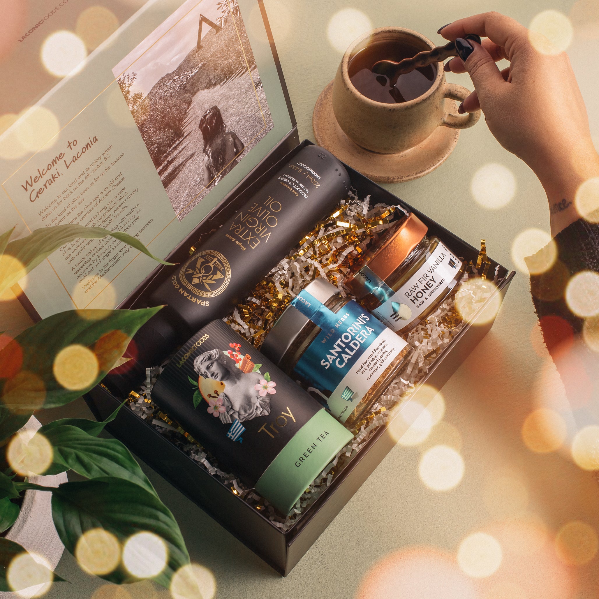 Lifestyle scene showing an open Laconic Foods Greek gift box with olive oil, tea, raw honey, and spices next to a freshly brewed cup of tea