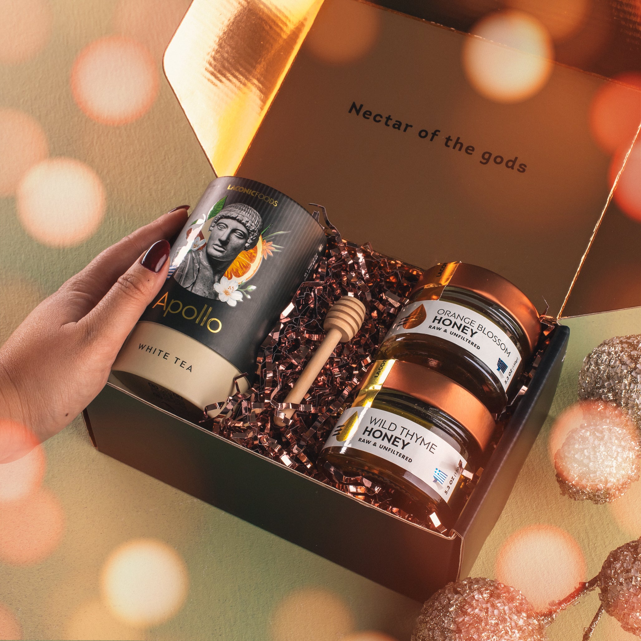 Lifestyle scene showing a hand opening a Laconic Foods Greek tea and honey gift box with Apollo white tea, raw honey jars, and honey dipper