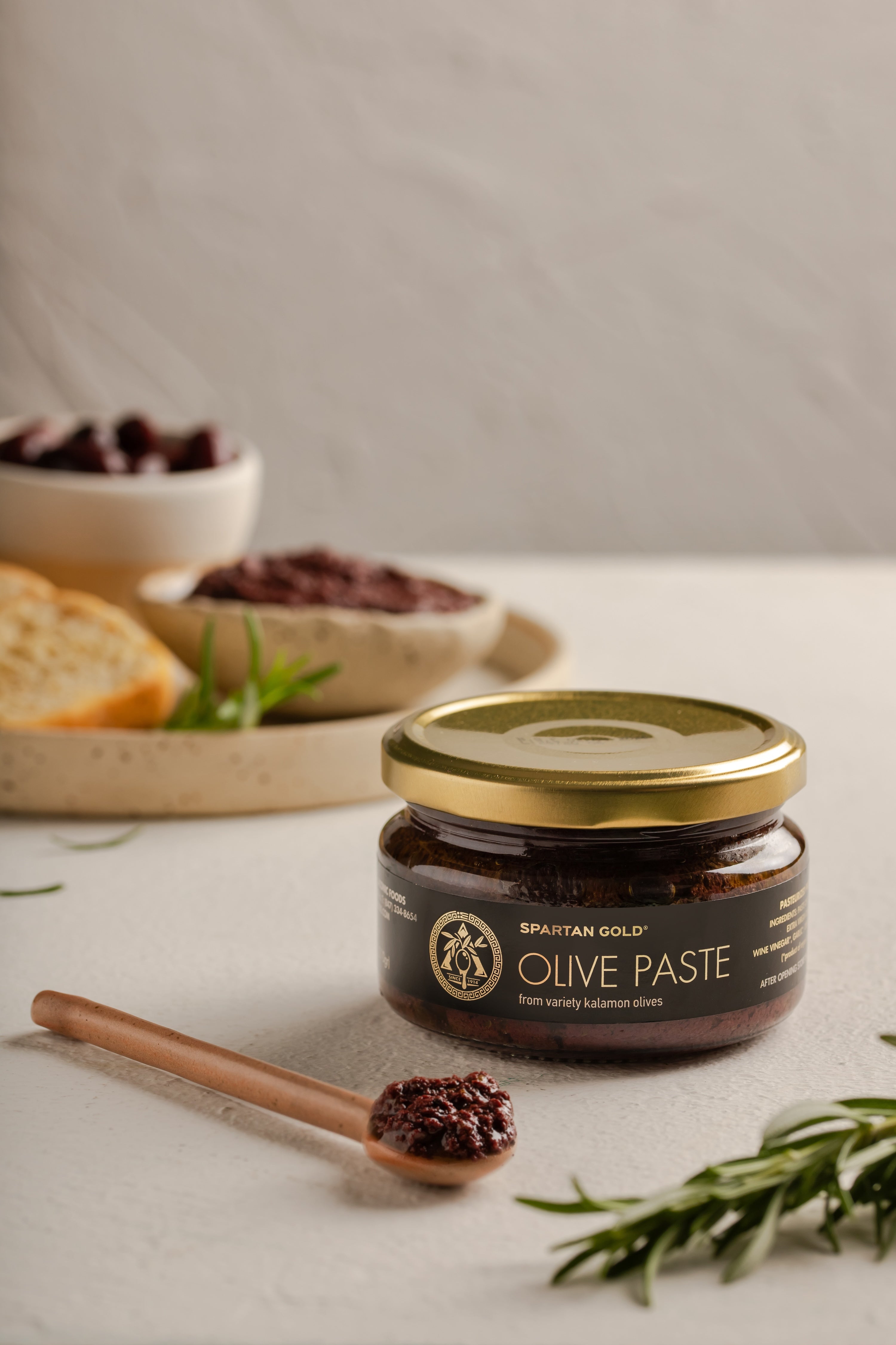 Glass jar of Laconic Foods Kalamata Olive Paste with wooden spoon, fresh rosemary, and bread on a white countertop