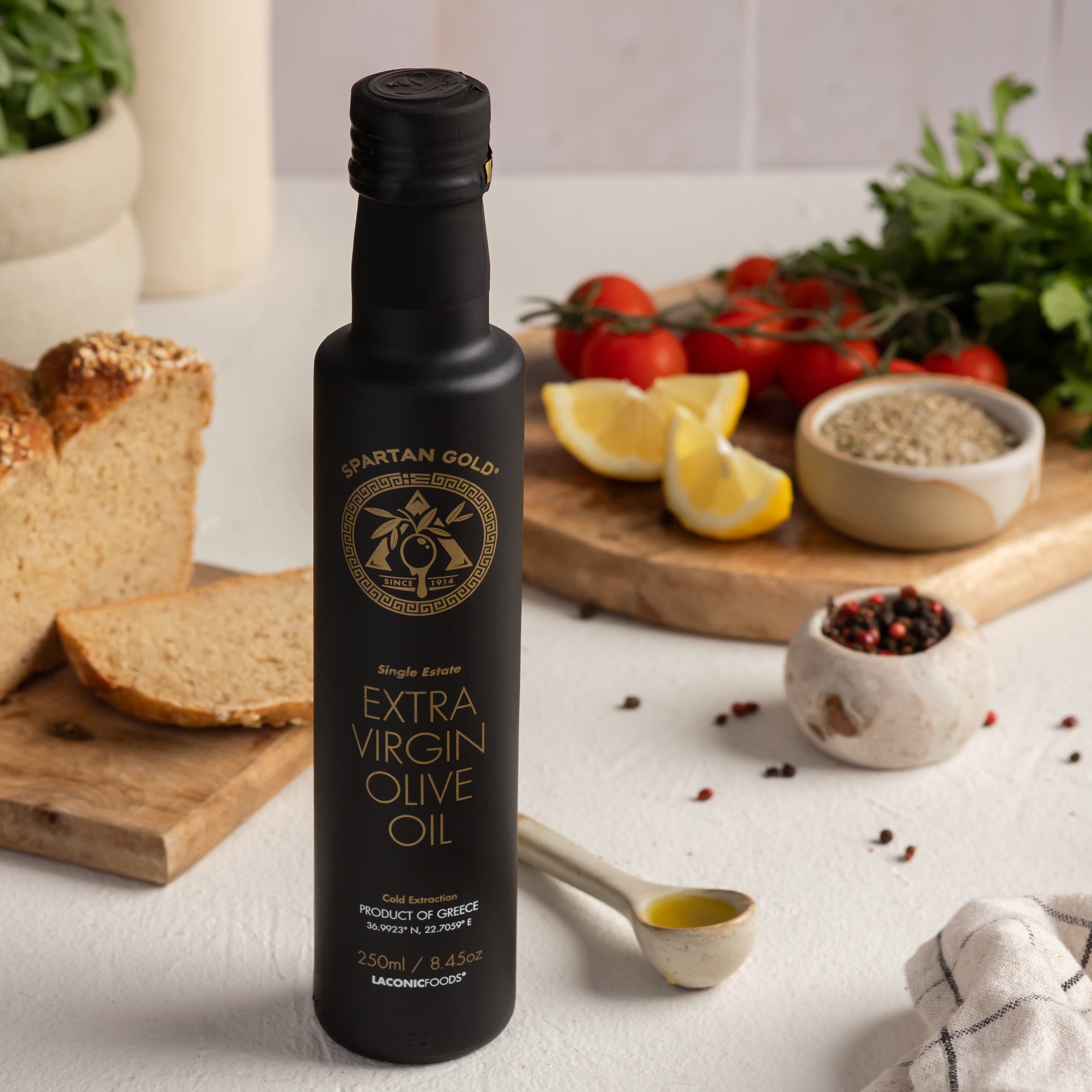 250ml bottle of Laconic Foods Extra Virgin Olive Oil with bread, lemon, tomatoes, herbs, and pepper in a Mediterranean kitchen setting