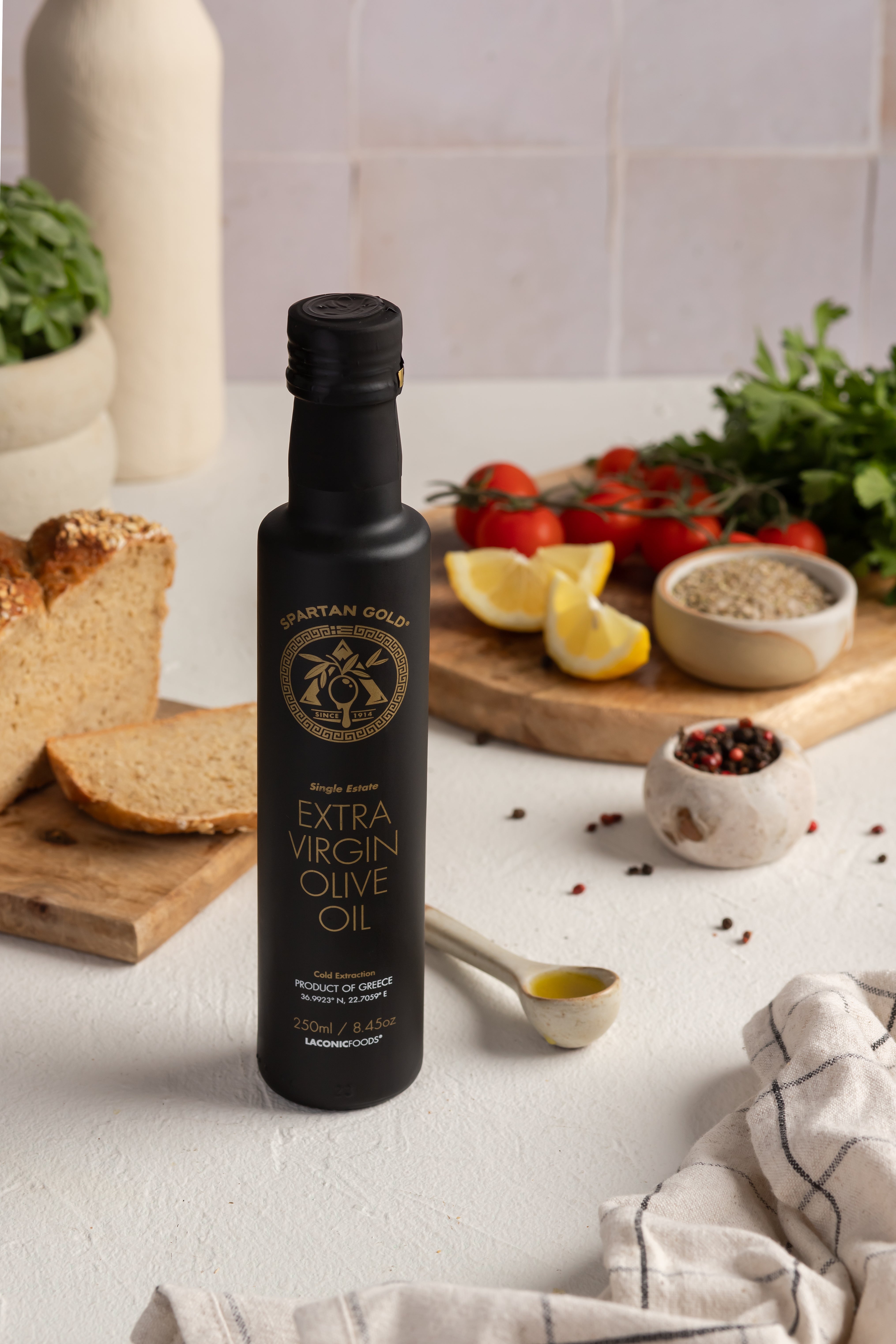 250ml bottle of Laconic Foods Extra Virgin Olive Oil with bread, lemon, herbs, and cracked pepper in a Mediterranean kitchen setting