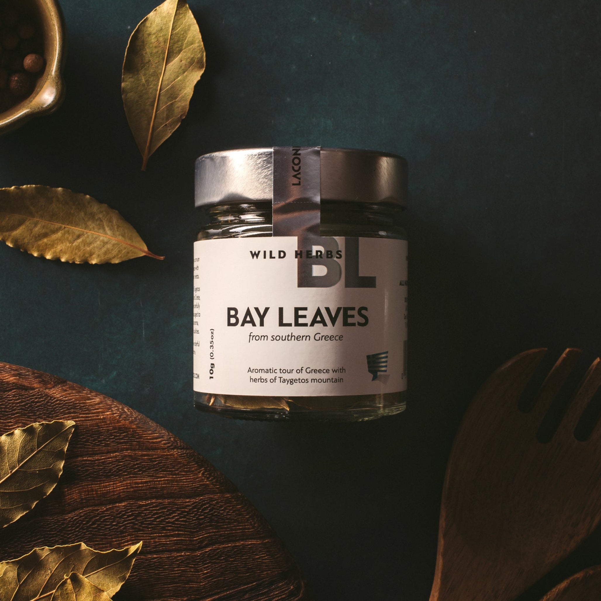 Laconic Foods Greek bay leaves wild herbs in a glass jar with dried bay leaves, wooden utensils, and rustic styling.