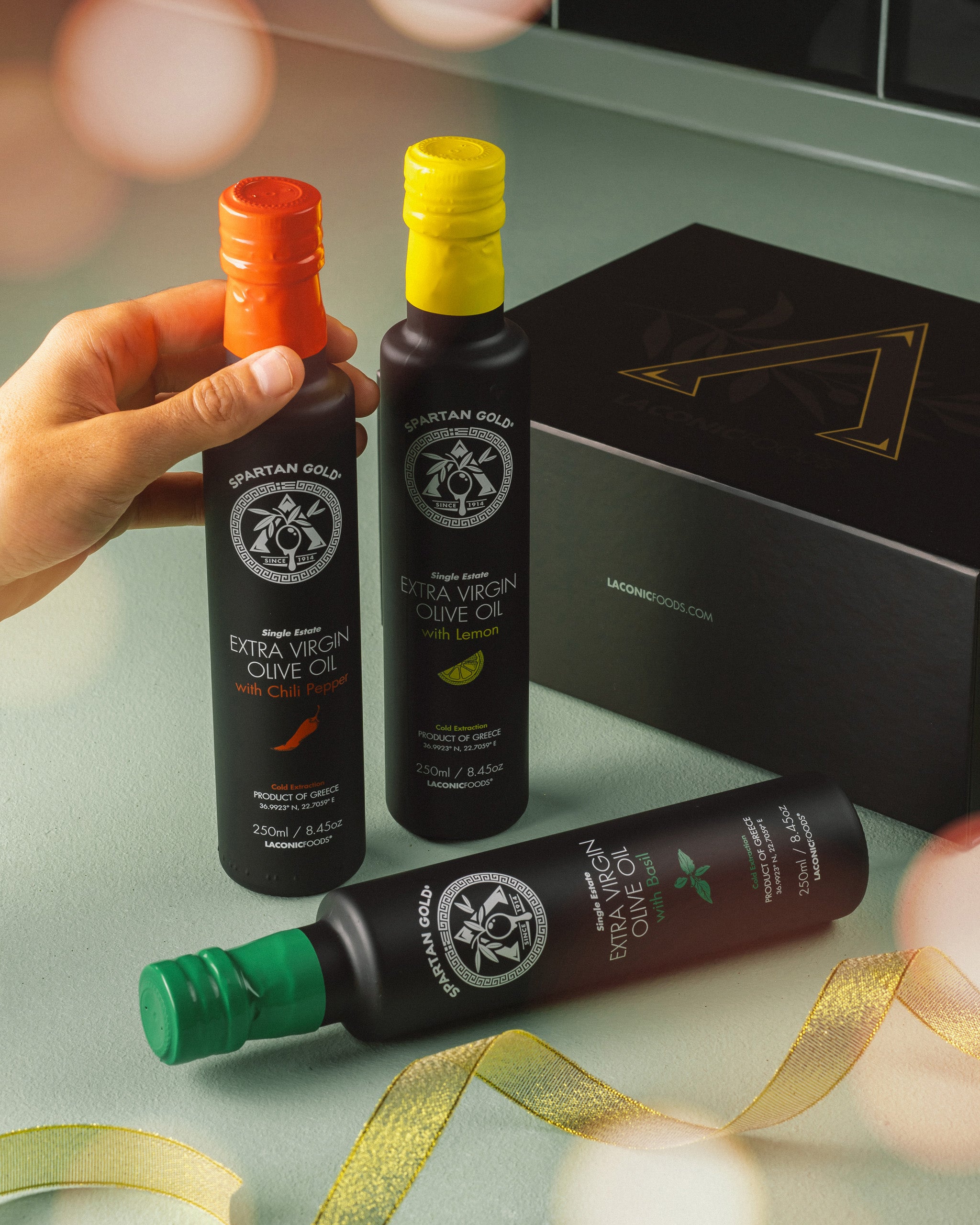 Hand holding Laconic Foods infused olive oil bottles with gift box in background, styled with ribbon.