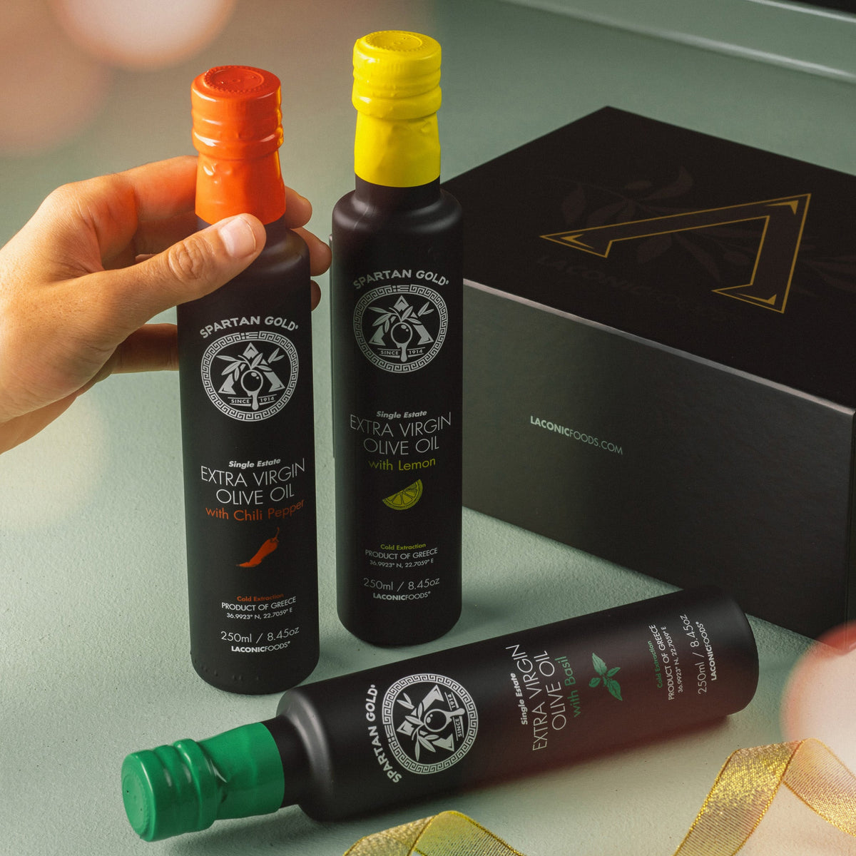 Hand holding Laconic Foods infused olive oil bottles with gift box in background, styled with ribbon.