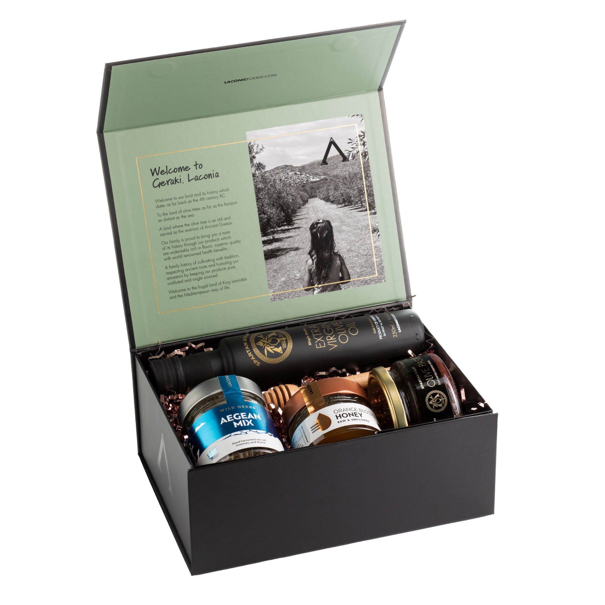 Open Laconic Foods gift box with extra virgin olive oil, raw honey, olive paste, and Aegean spice blend, with printed grove photo and family story inside lid