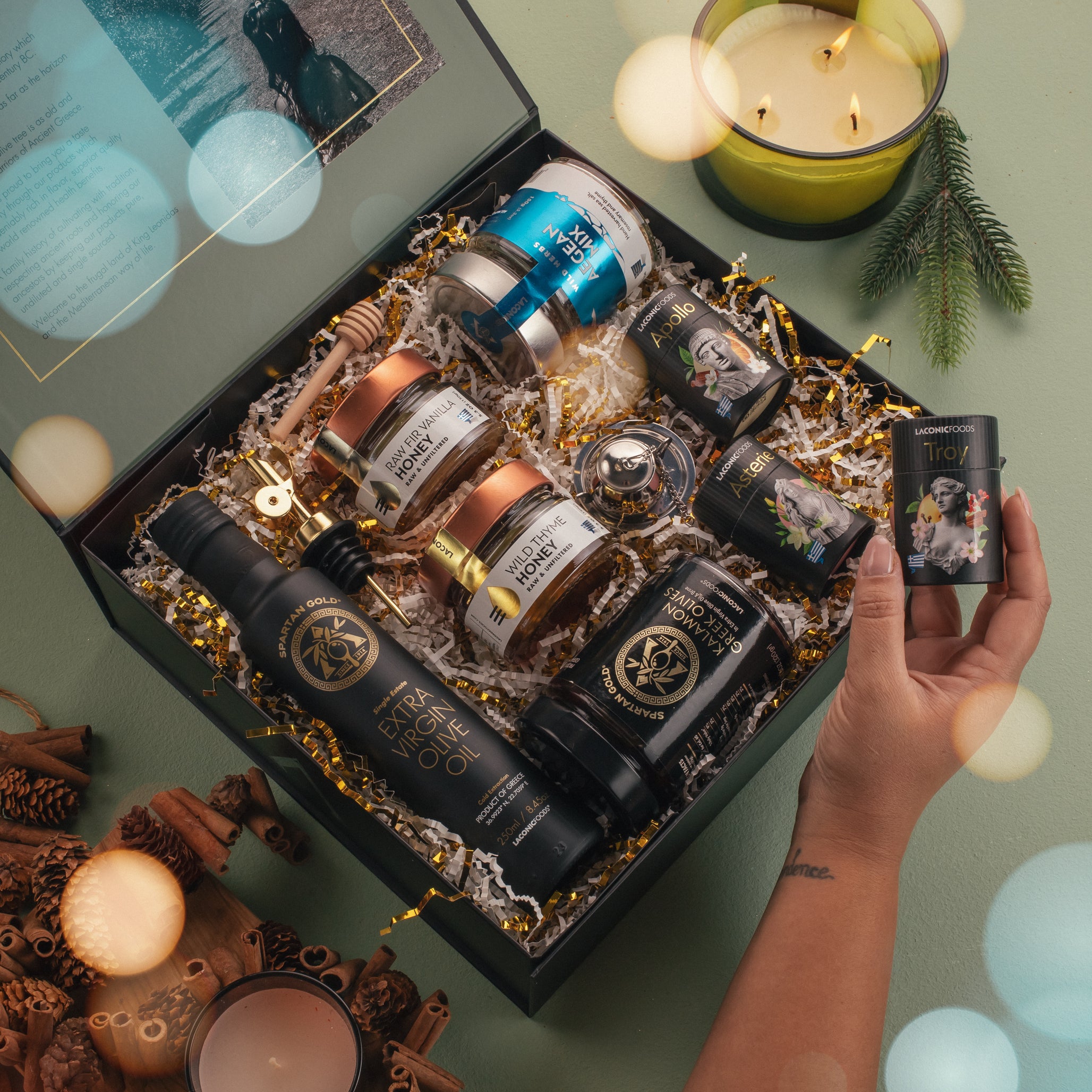 Lifestyle photo of the Laconic Foods Premium Greek Gift Box opened with teas, honeys, olives, EVOO, Aegean spice mix, and tea infuser, styled with candles and holiday dΓ©cor.