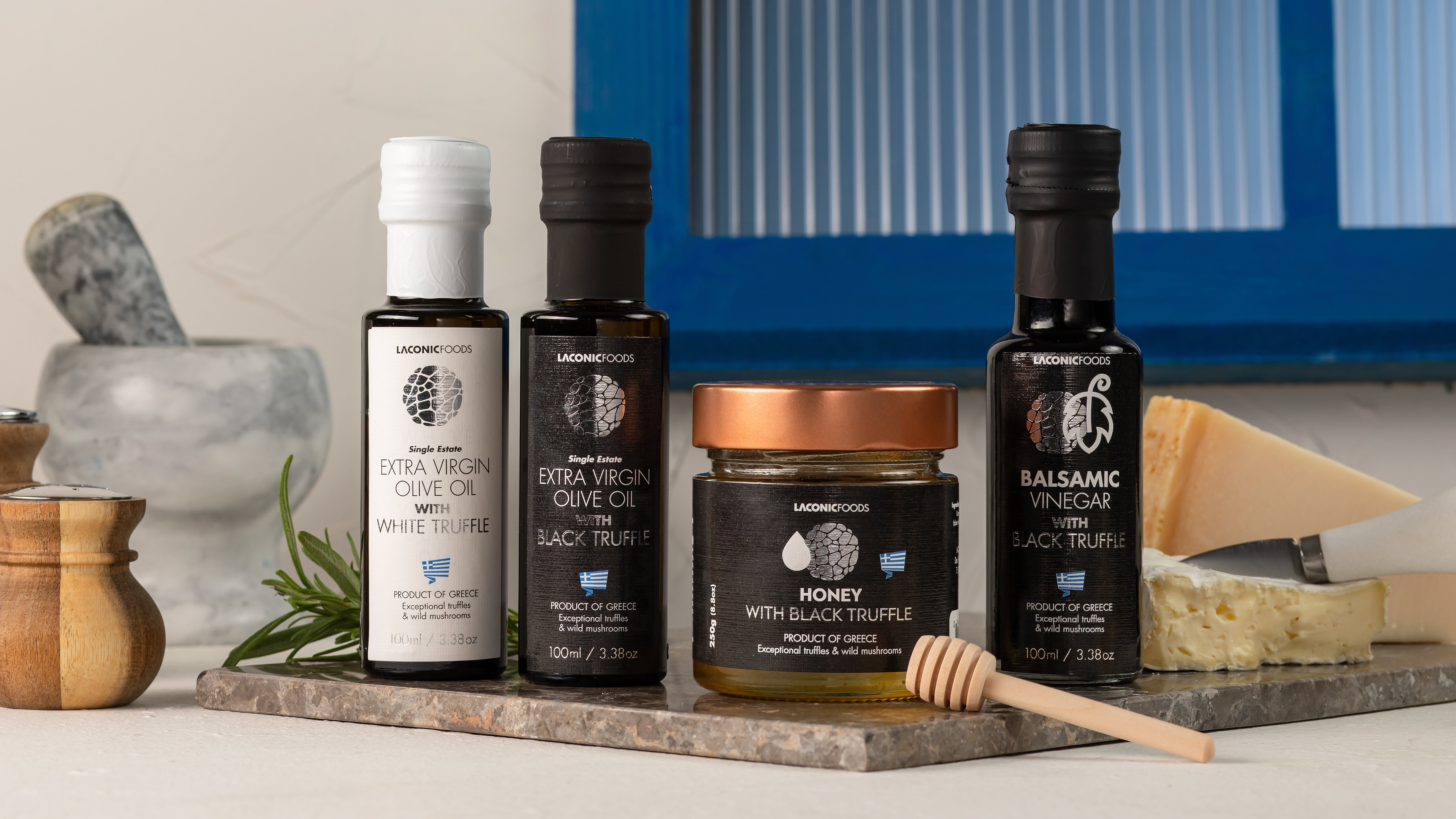 Laconic Foods truffle collection featuring White Truffle EVOO, Black Truffle EVOO, Black Truffle Honey, and Black Truffle Balsamic Vinegar arranged with cheese and herbs.