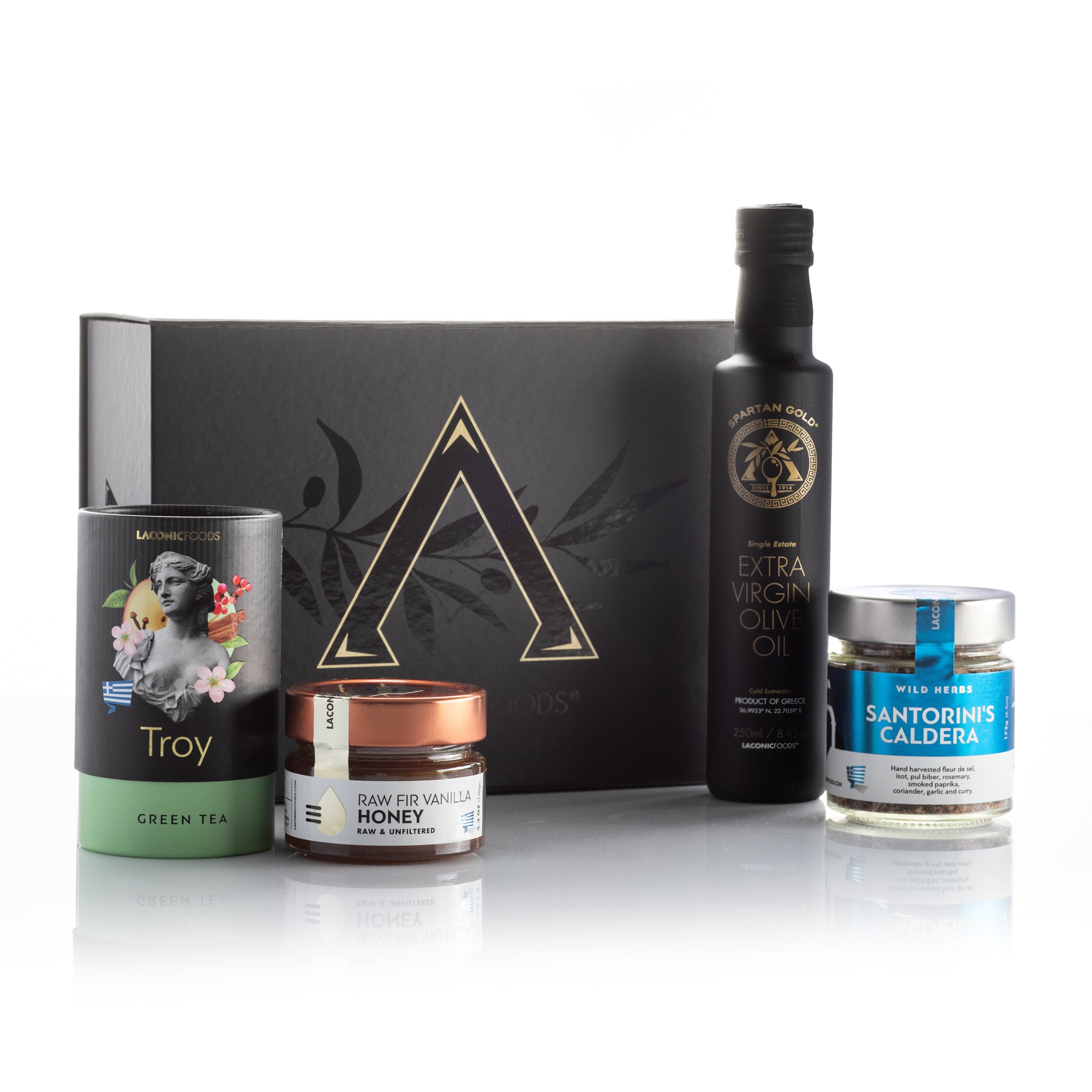 Laconic Foods Greek gift set displayed with extra virgin olive oil bottle, premium loose leaf tea, raw honey, and Aegean herb blend beside a black gift box