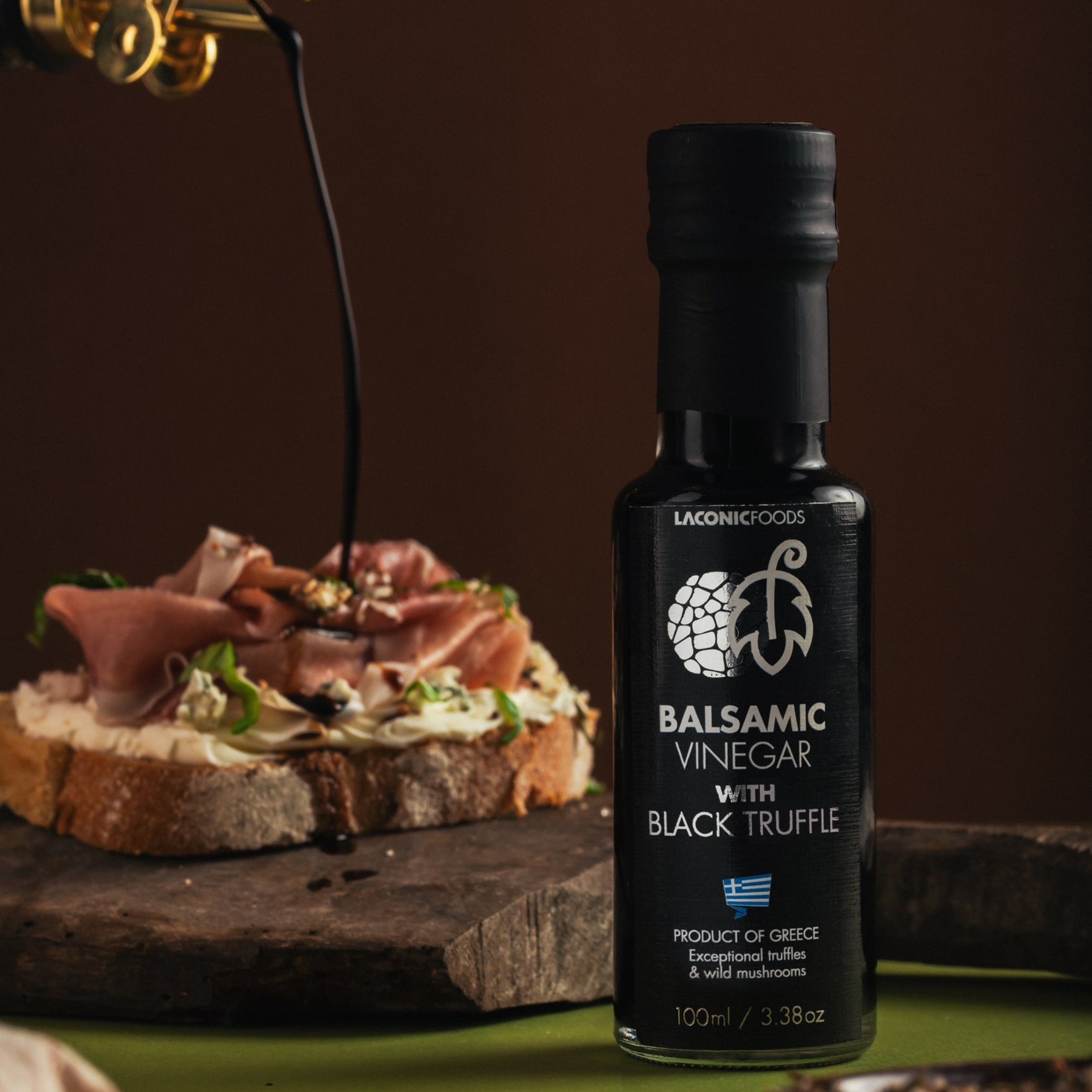 Laconic Foods Balsamic Vinegar with Black Truffle displayed on a rustic board with balsamic being poured over artisan bread.