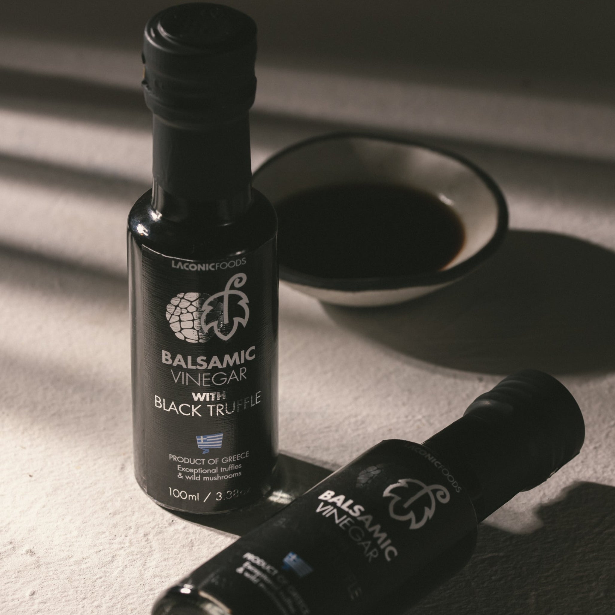 Two bottles of Laconic Foods Balsamic Vinegar with Black Truffle on a textured surface with dramatic shadows and a bowl of balsamic.