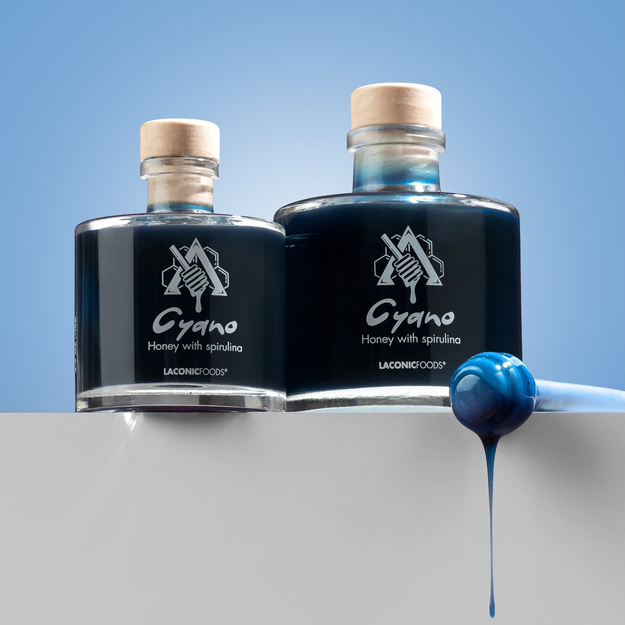 Two glass bottles of Cyano blue honey with spirulina and a honey dipper — Laconic Foods product shot.