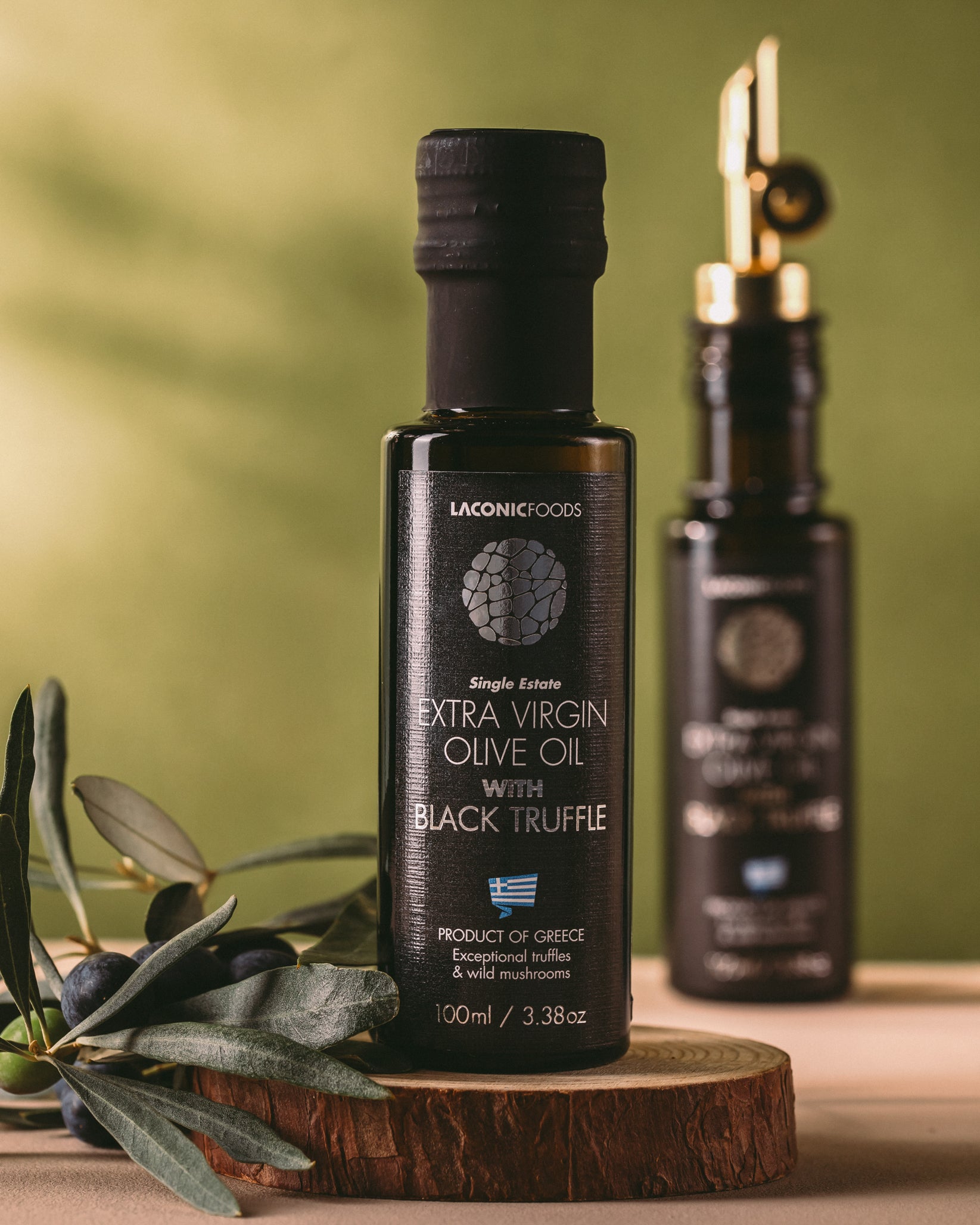 Bottle of Laconic Foods Extra Virgin Olive Oil with Black Truffle displayed on a wooden board with fresh olives and olive leaves.