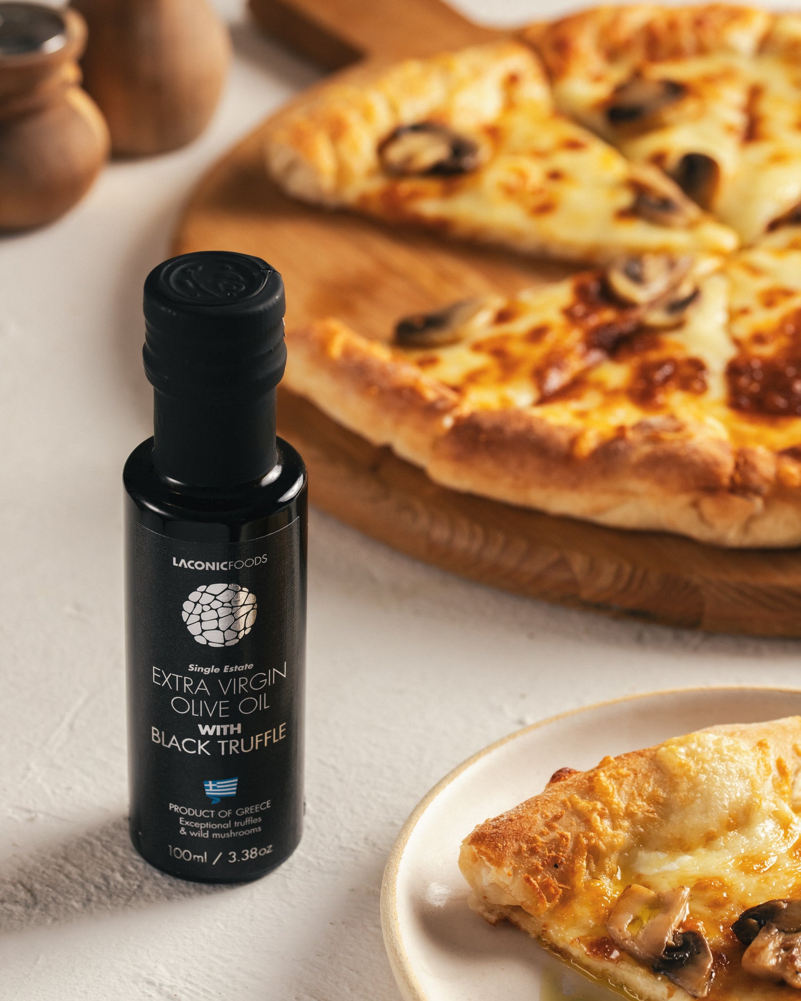 Laconic Foods Extra Virgin Olive Oil with Black Truffle placed beside a mushroom pizza slice on a plate and wooden pizza board.
