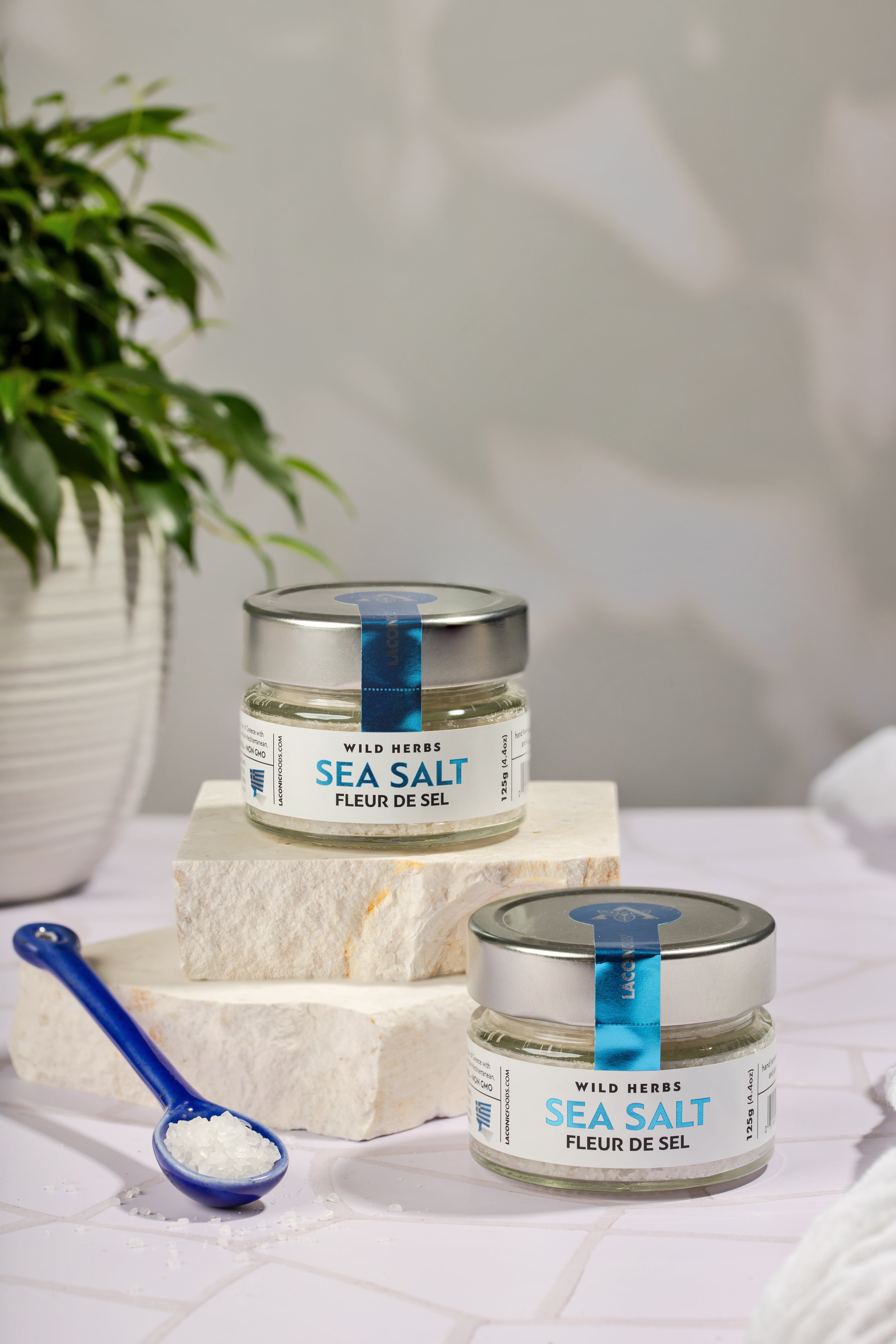Two jars of Laconic Foods Fleur de Sel Sea Salt with ceramic spoon and Greek stone background
