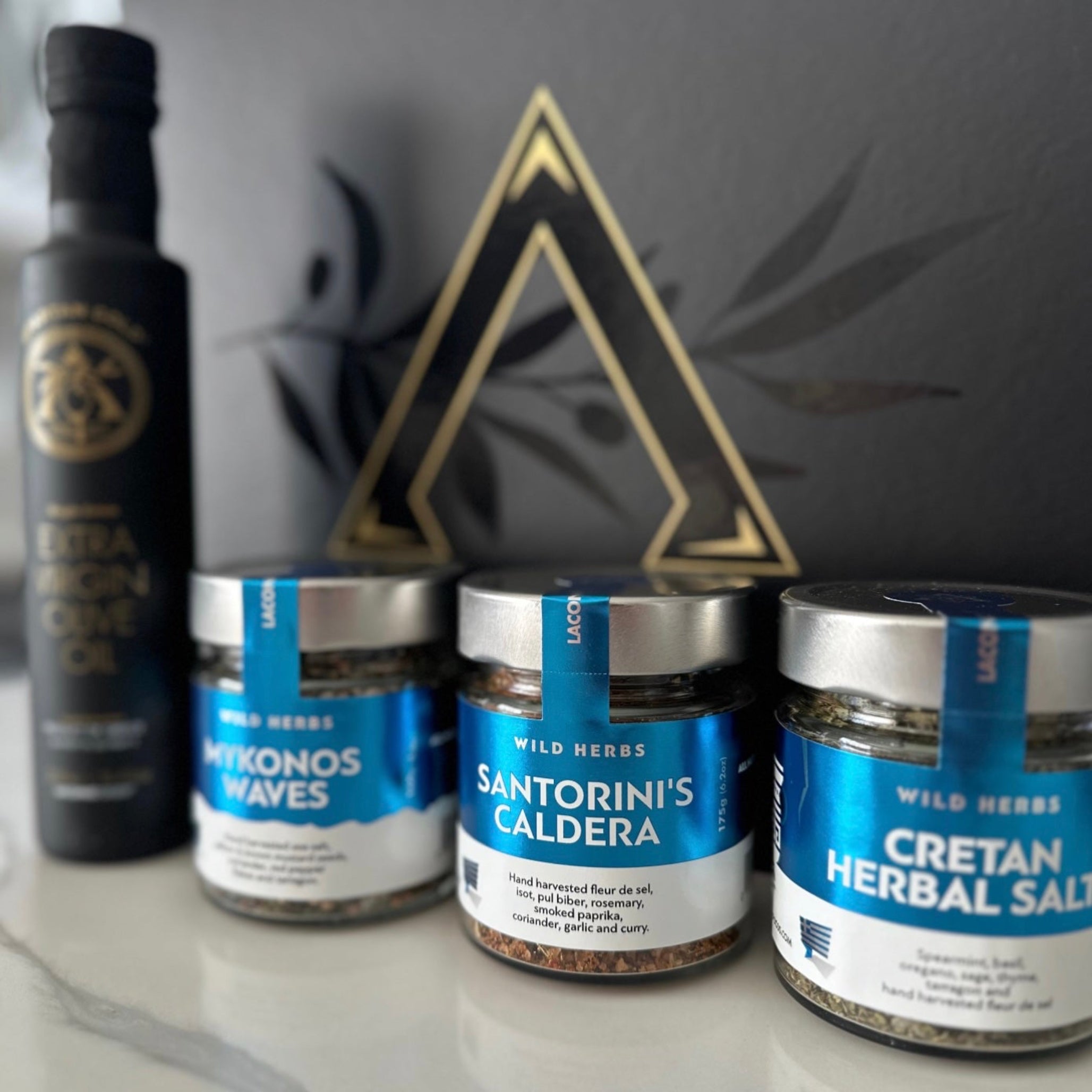 Close-up of Laconic Foods Mykonos Waves, Santorini’s Caldera, and Cretan Herbal Salt jars with extra virgin olive oil bottle