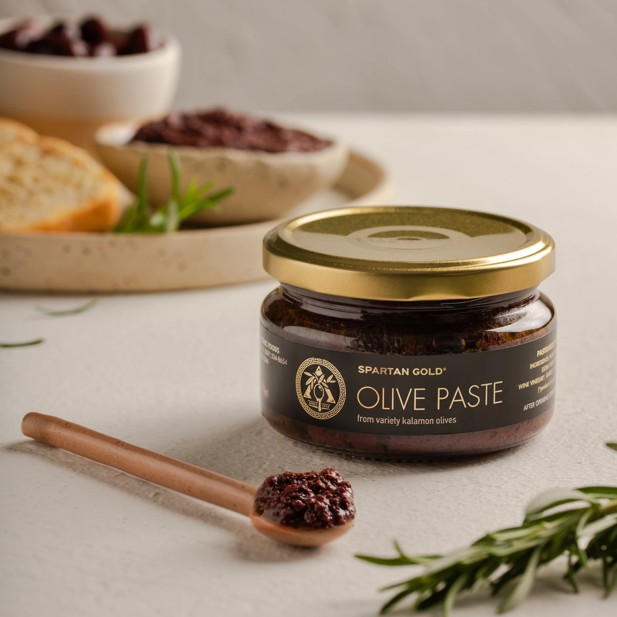 Glass jar of Laconic Foods Kalamata Olive Paste with wooden spoon, fresh rosemary, and bread on a white countertop