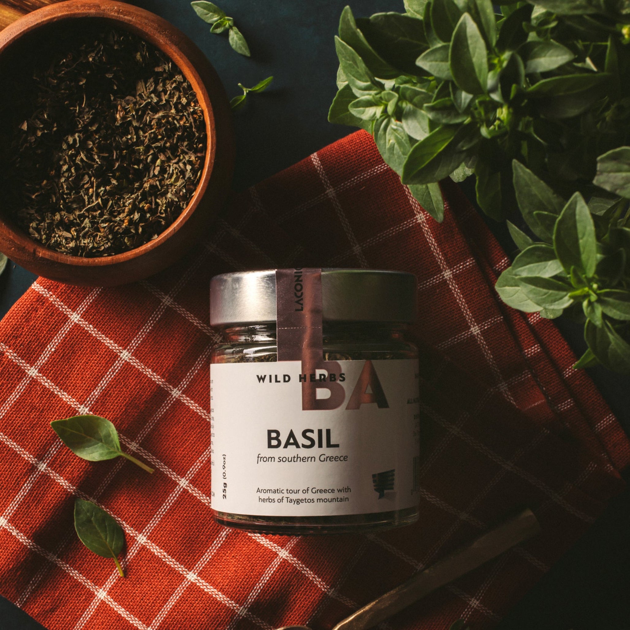 Laconic Foods Greek basil wild herbs in a glass jar with fresh basil leaves, dried basil, a spoon, and red kitchen cloth.