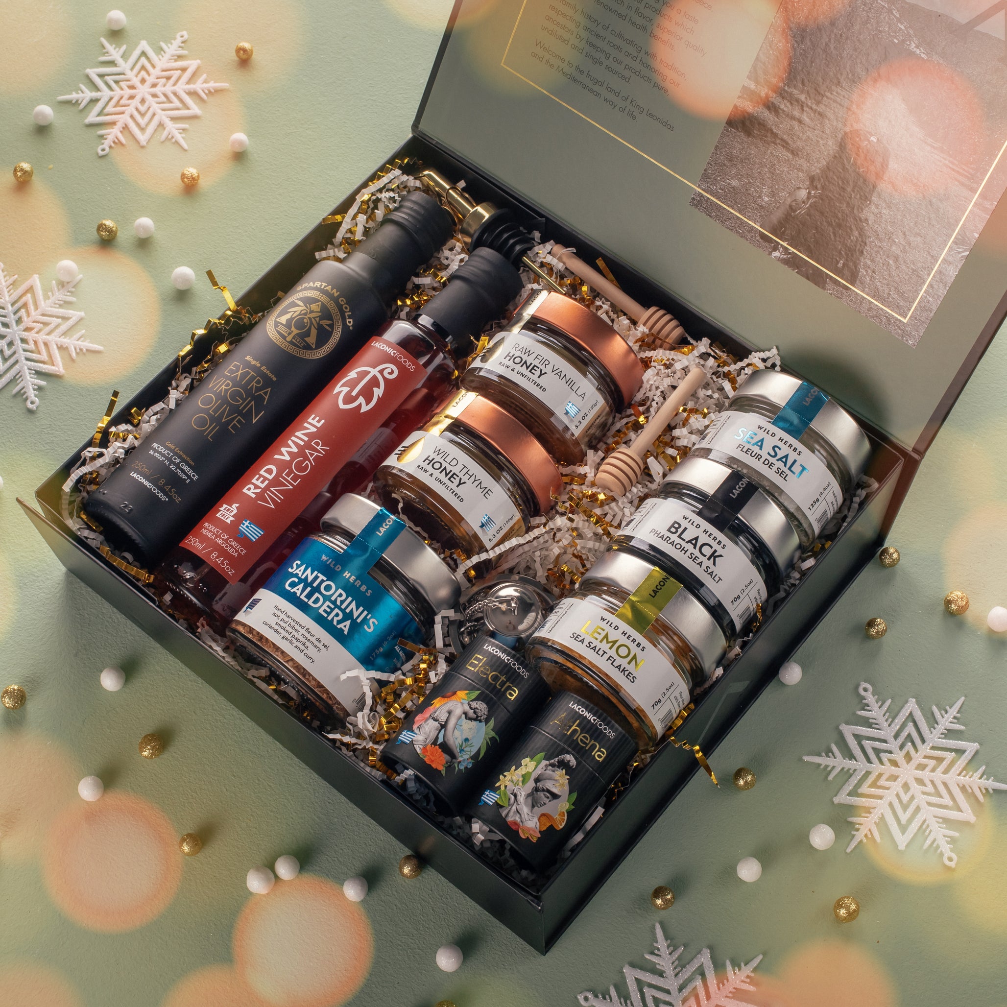 Festive holiday presentation of the Laconic Foods Luxury Greek Gift Box opened on a decorated background with snowflakes, showcasing olive oil, red wine vinegar, honeys, salts, teas, and Santorini spice blend.