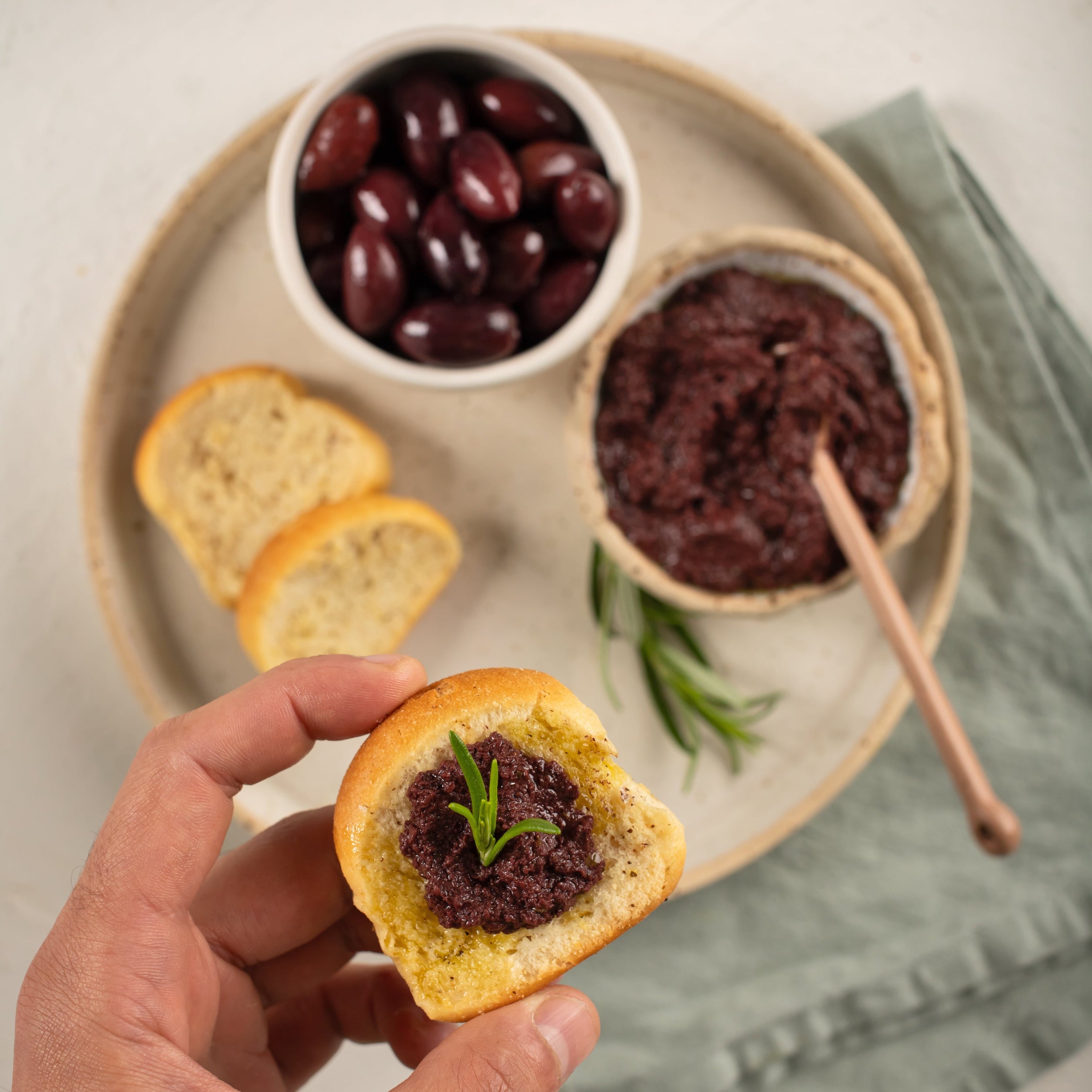 Kalamata Olive Paste spread on toasted bread, held in hand with rosemary garnish