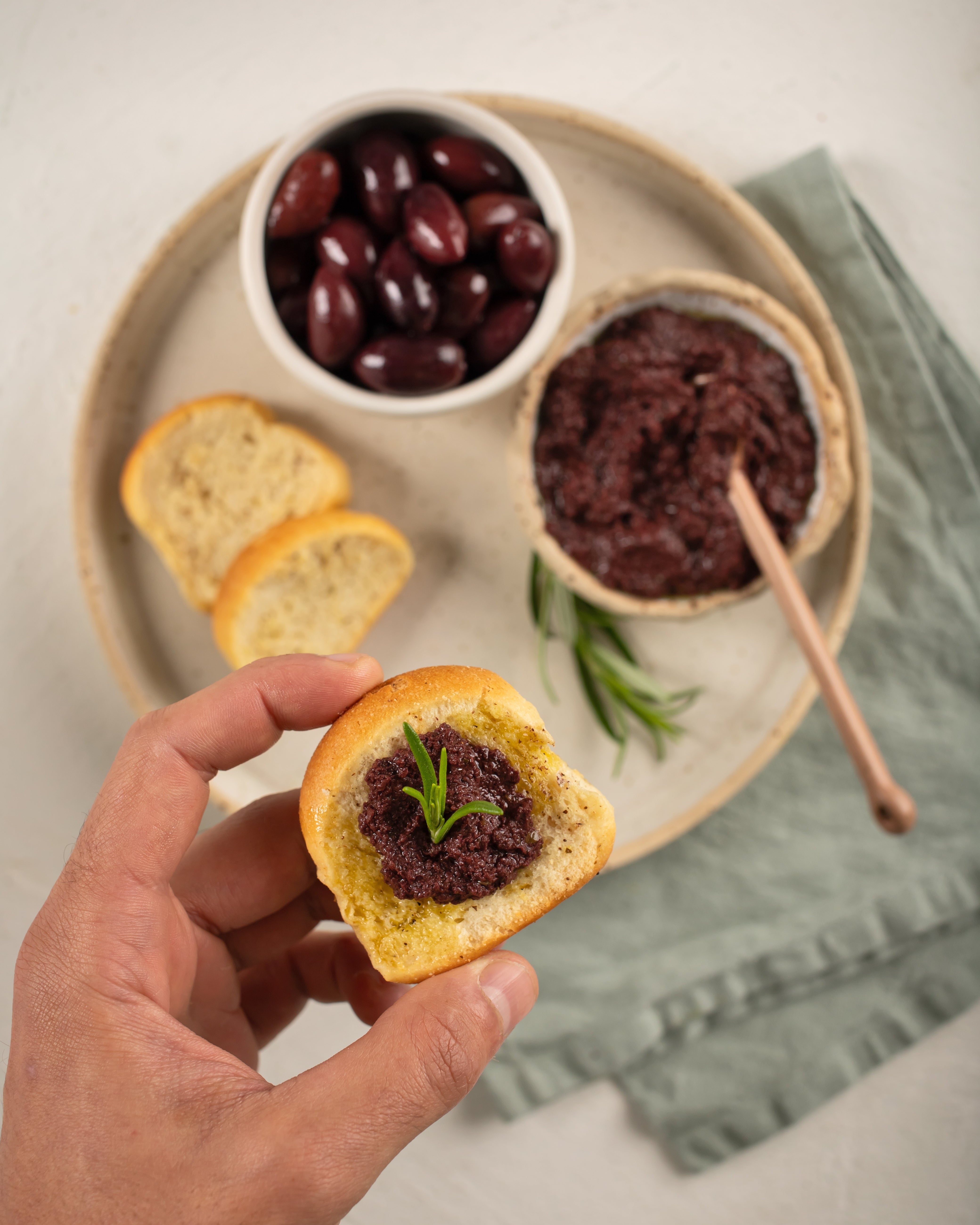 Kalamata Olive Paste spread on toasted bread, held in hand with rosemary garnish