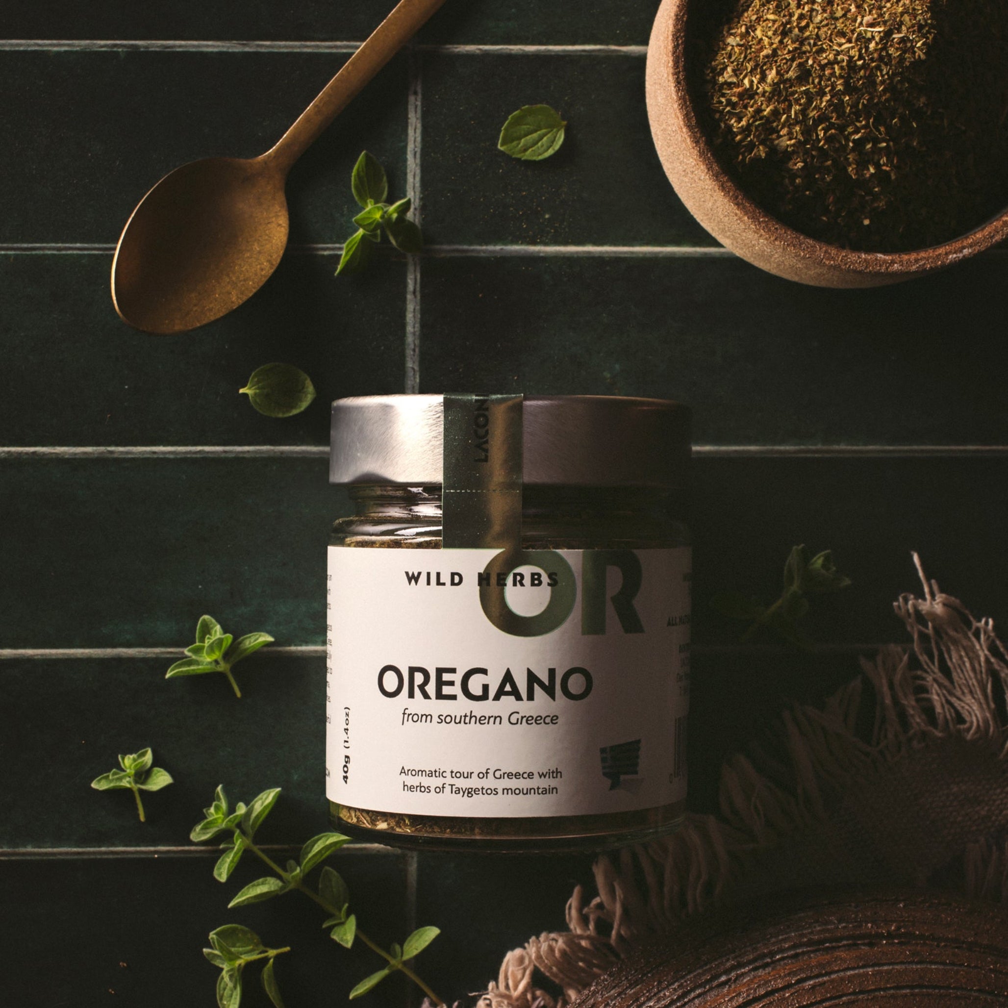 Laconic Foods Greek oregano wild herbs in a glass jar with fresh oregano leaves, dried oregano, a spoon, and rustic styling.