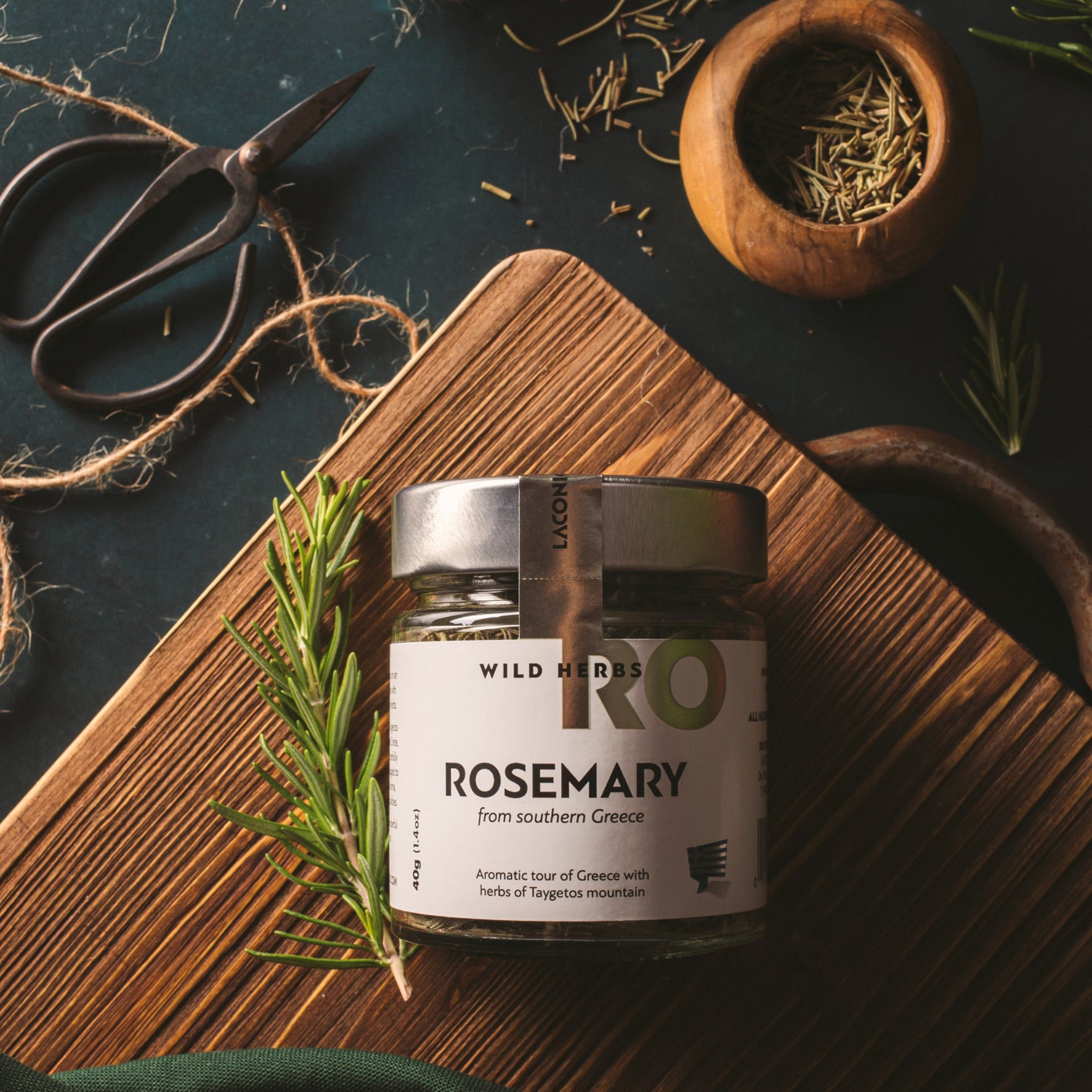 Laconic Foods Greek rosemary wild herbs in a glass jar on a wooden board with fresh rosemary, dried rosemary, herb scissors, and rustic styling.