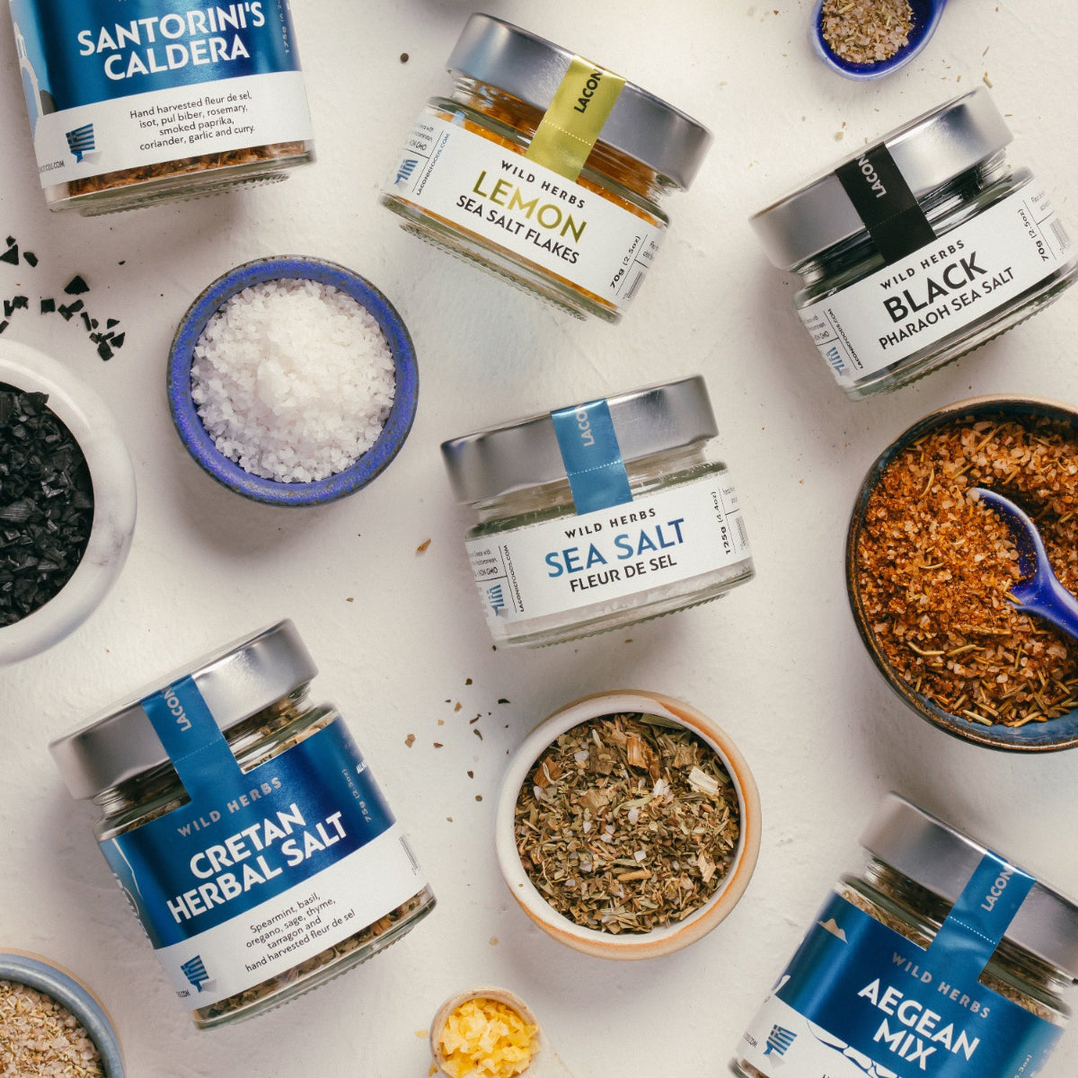 Flat lay of Laconic Foods Greek spices and sea salts, including Santorini Caldera blend, Mykonos Waves, Cretan herbal salt, fleur de sel, lemon salt flakes, and black sea salt.