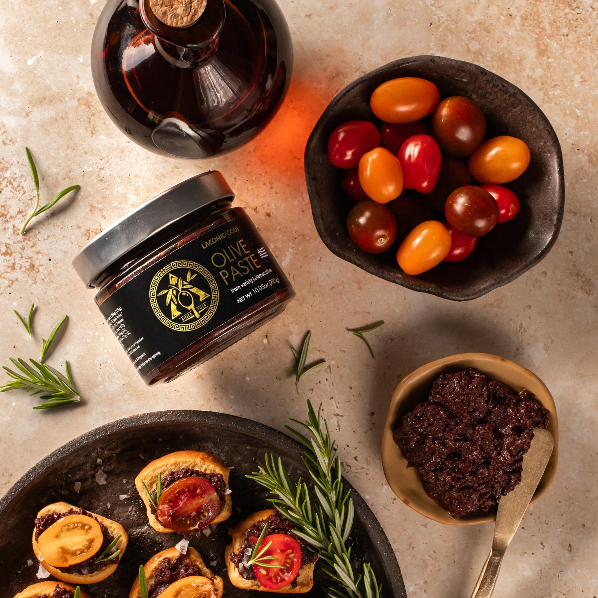 Laconic Foods Kalamata olive paste with crostini, cherry tomatoes, rosemary, and olive oil on a rustic stone surface.