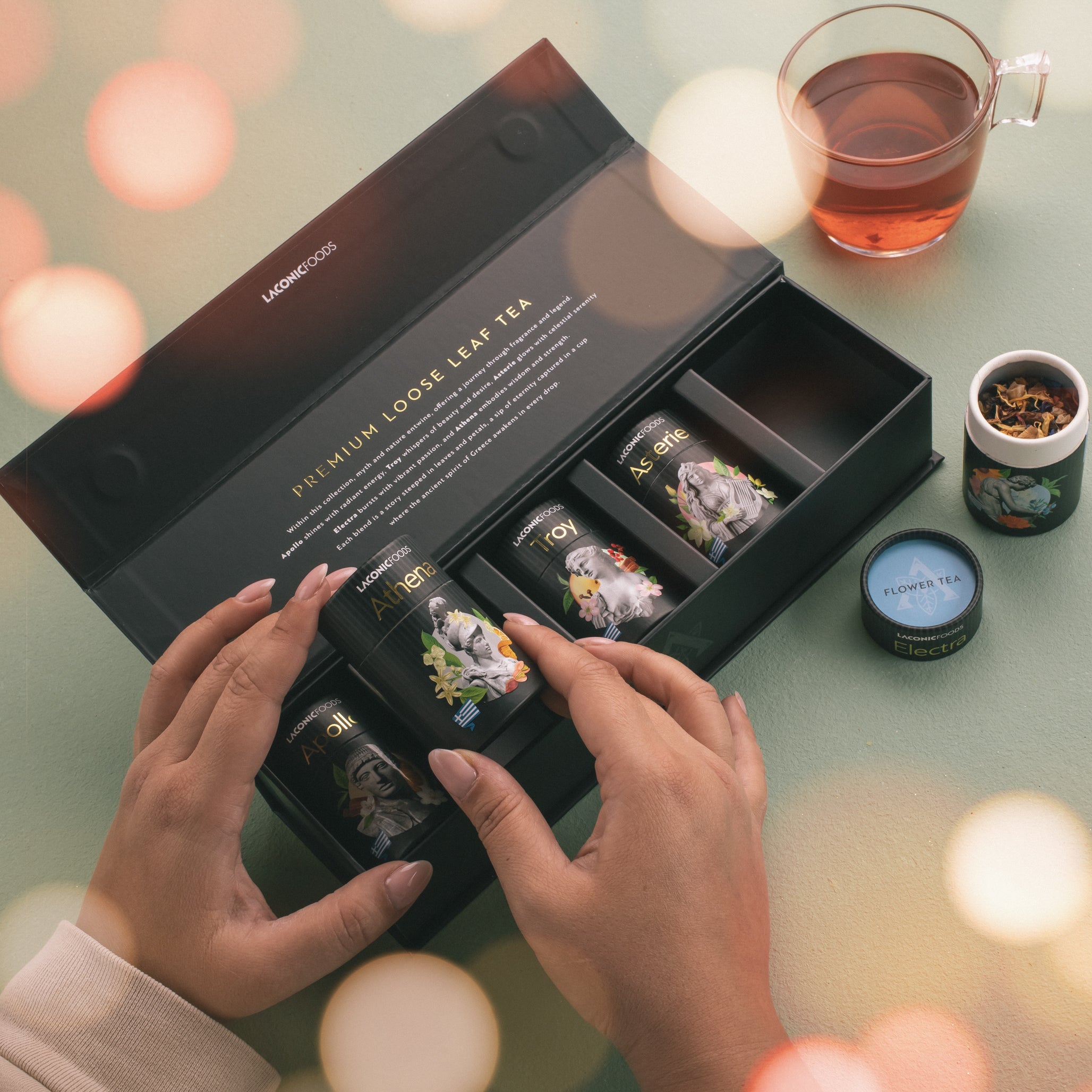 Lifestyle image of Laconic Foods mini loose leaf tea gift set with hands opening a luxury box, tea tins, loose tea leaves, and brewed tea