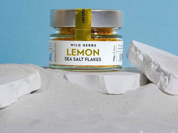 Lemon Sea Salt Flakes 70g jar by Laconic Foods displayed in white sand with blue background