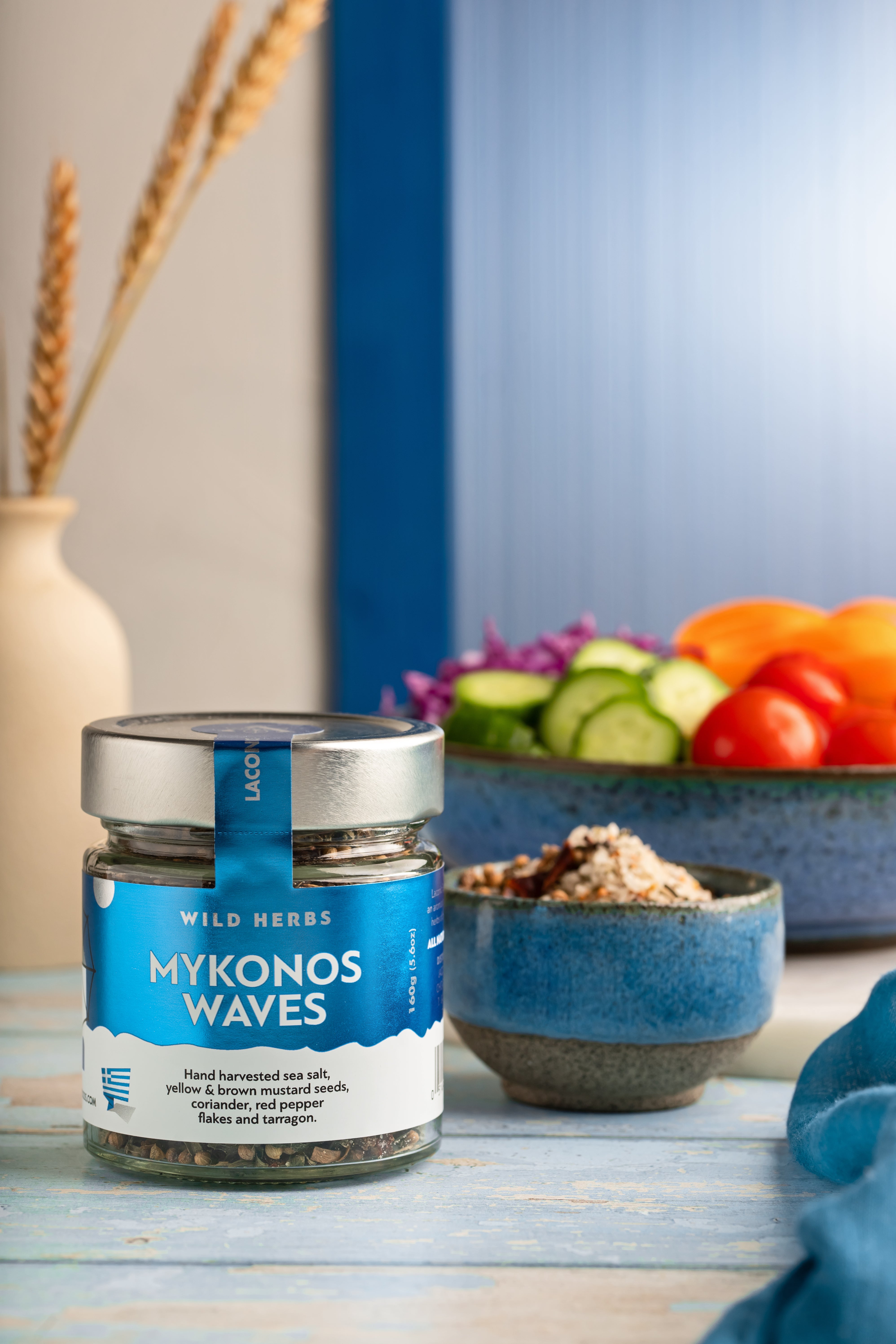 Mykonos Waves Greek Spice Blend styled beside a salad bowl with cucumbers, tomatoes, and Mediterranean ingredients