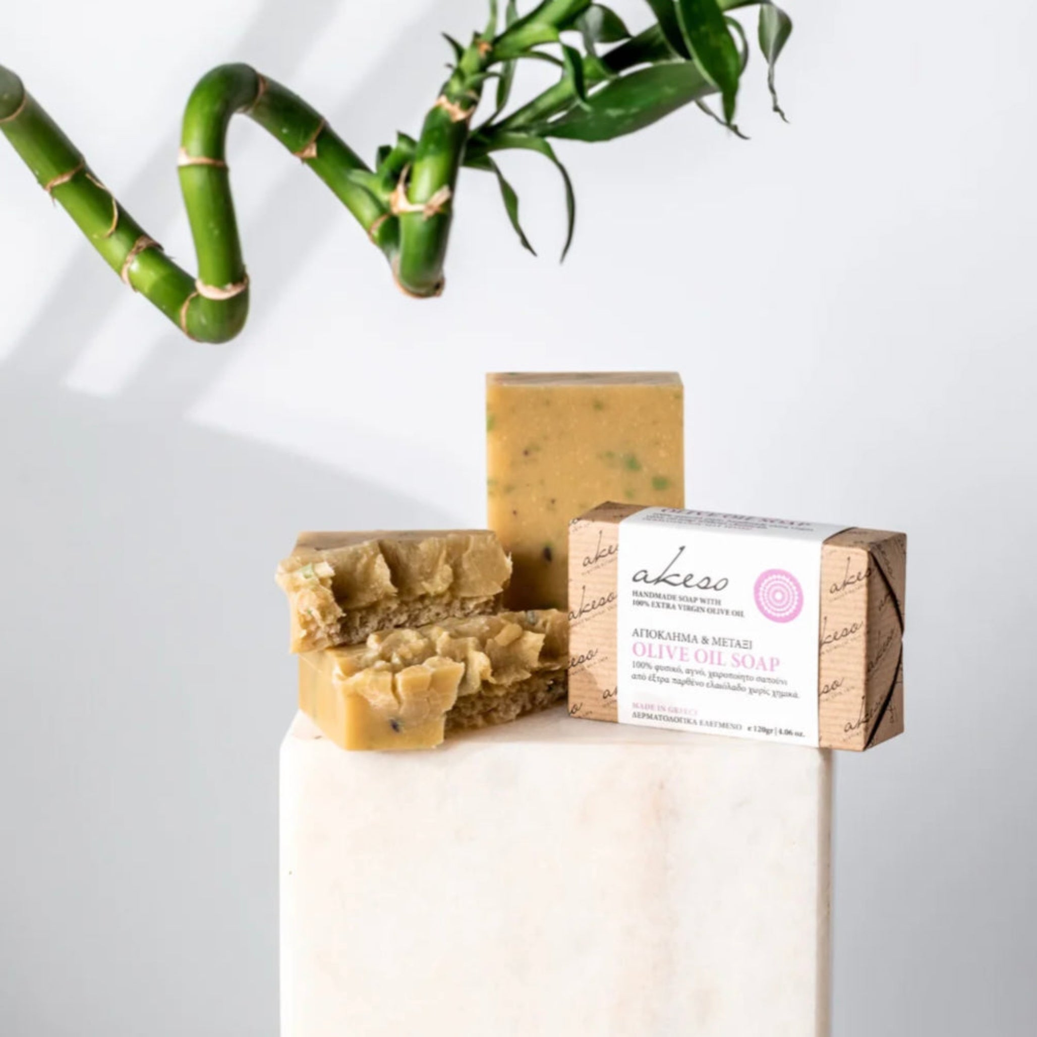 Handmade olive oil soap bars displayed with packaged Greek soap, bamboo leaves, and a white ceramic vase on a clean light background.