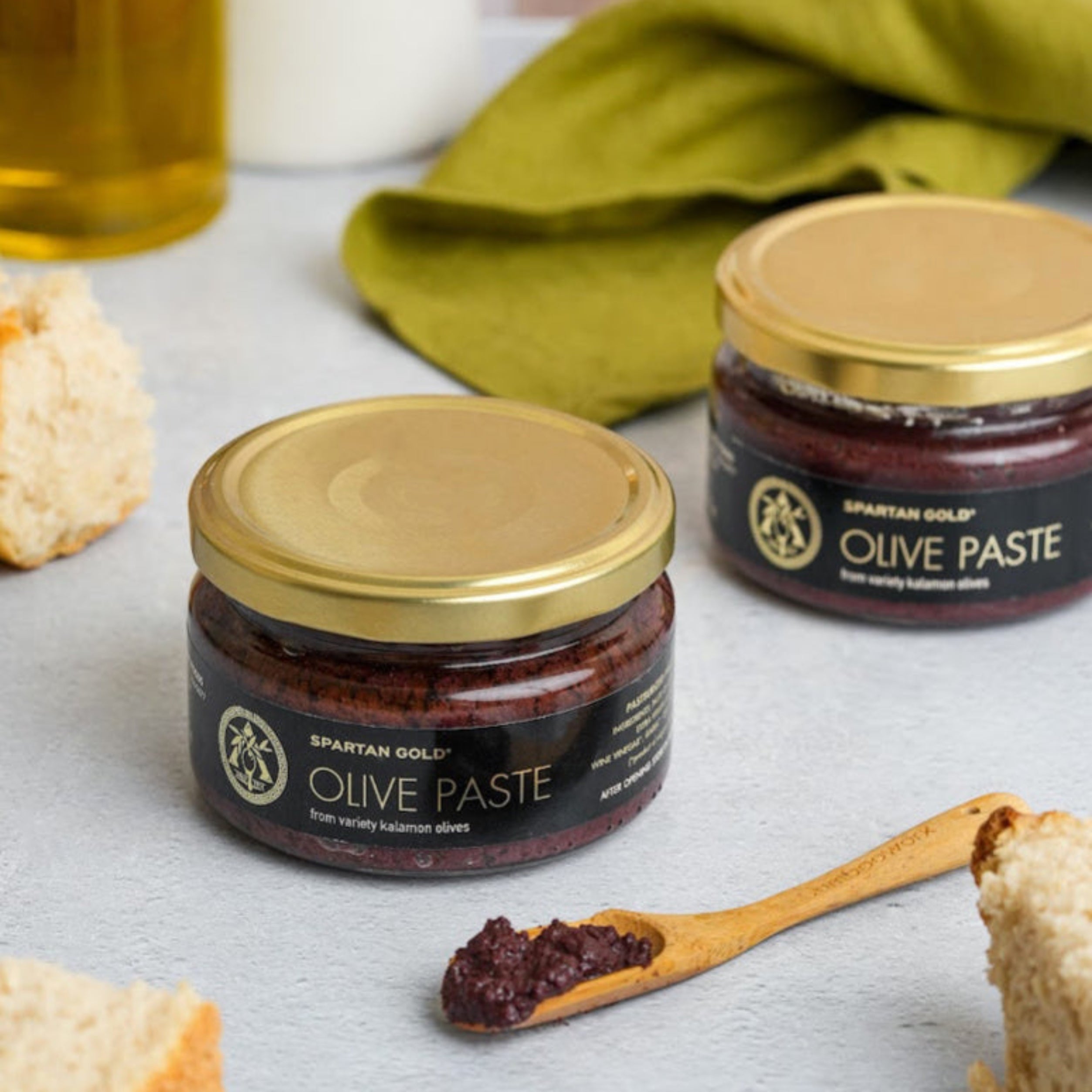 Two jars of Kalamata Olive Paste with spoonful served, surrounded by fresh bread and olive oil