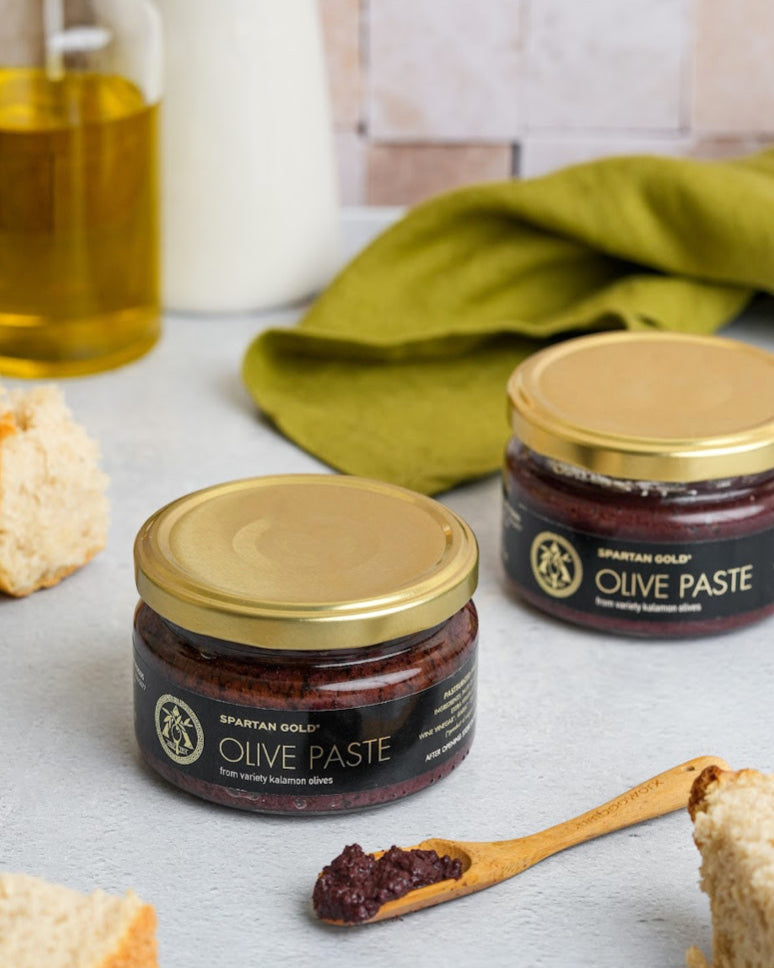 Two jars of Kalamata Olive Paste with spoonful served, surrounded by fresh bread and olive oil